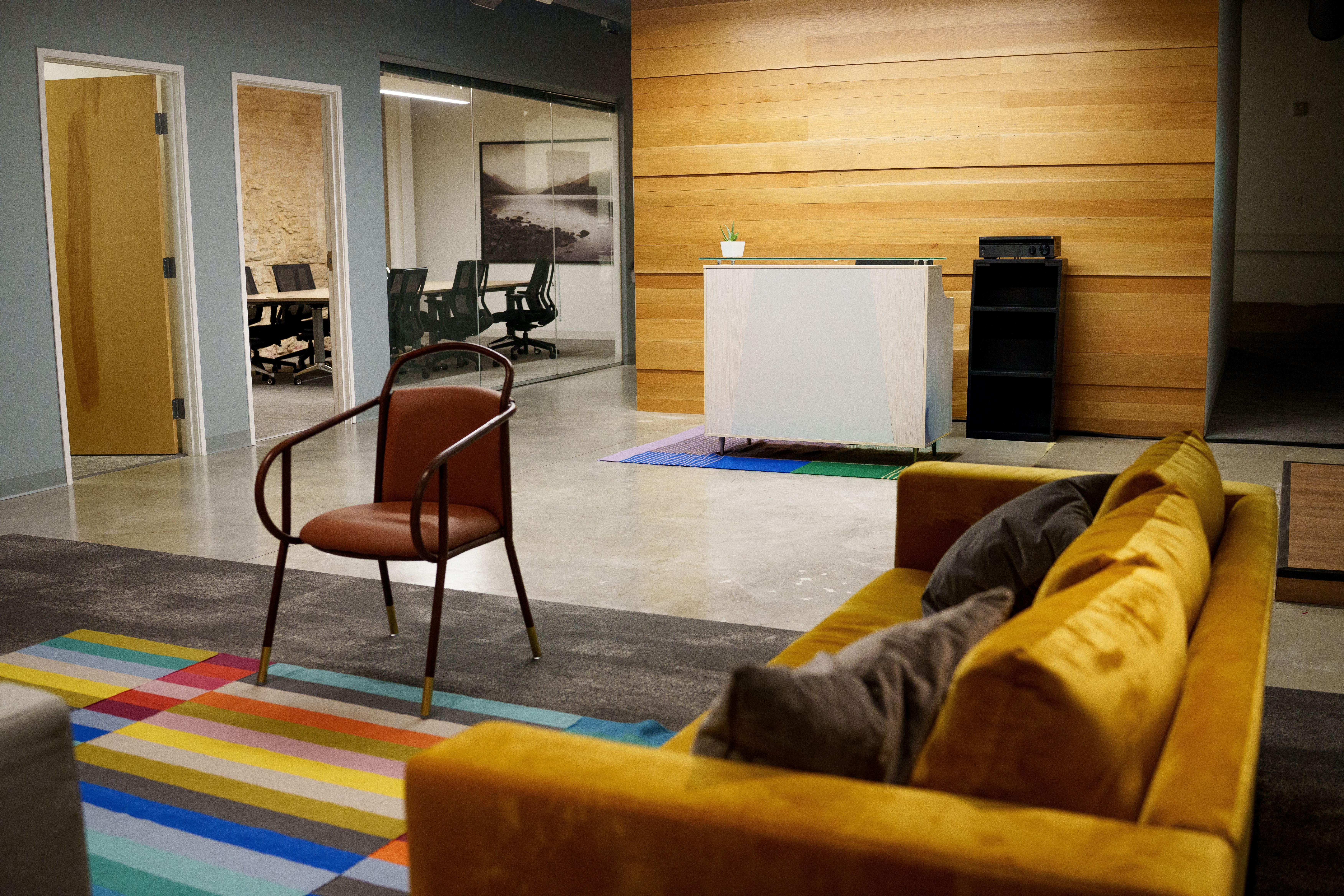 Modern office lobby with yellow sofa, rust-colored chair on striped rug, white reception desk on colorful mat, wood panel wall, and glass-walled meeting room with black chairs.