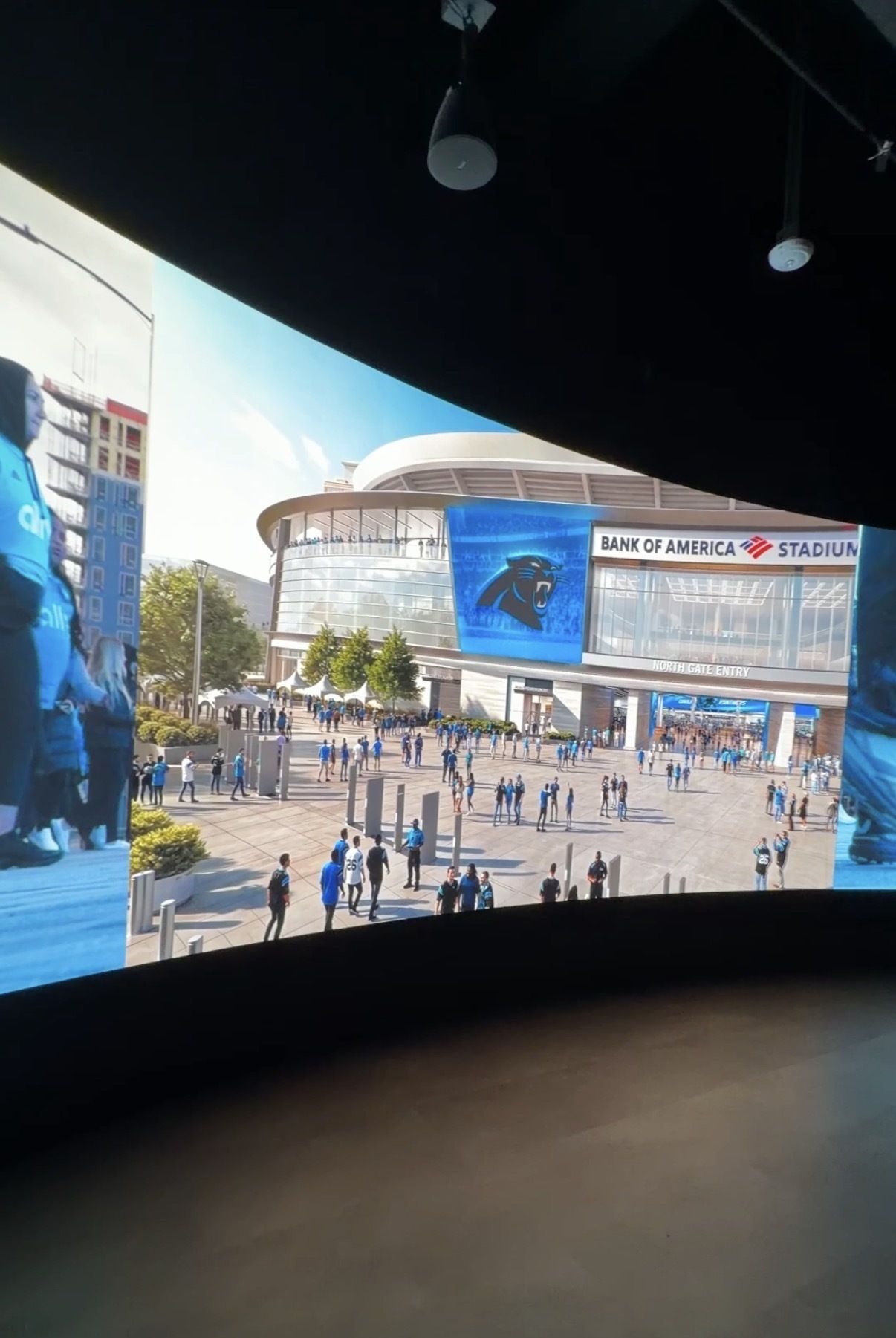 Outdoor view of Bank of America Stadium with a curved glass facade. A large blue digital screen shows the Carolina Panthers logo as crowds walk toward the North Gate Entry.