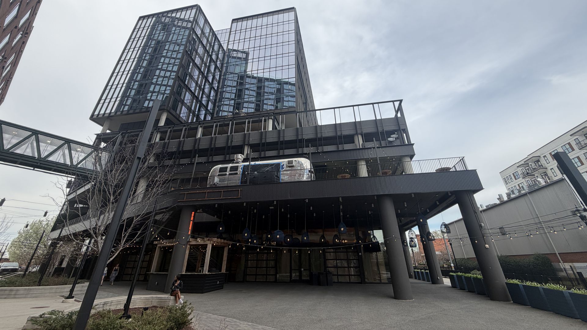 Low-angle view of a modern glass-and-steel building with an elevated platform; a sleek tram sits on the upper level, string lights along the entrance, and a person walking in the plaza.