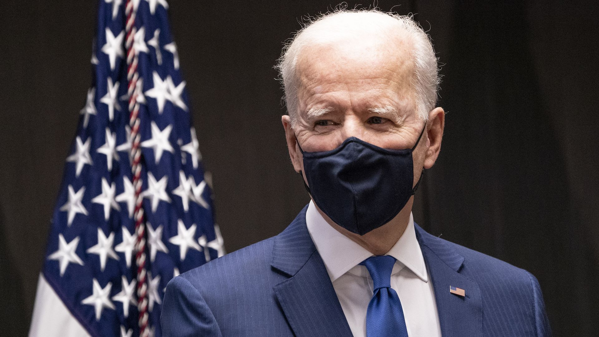 Picture of Biden wearing a black face mask and standing next to an American flag