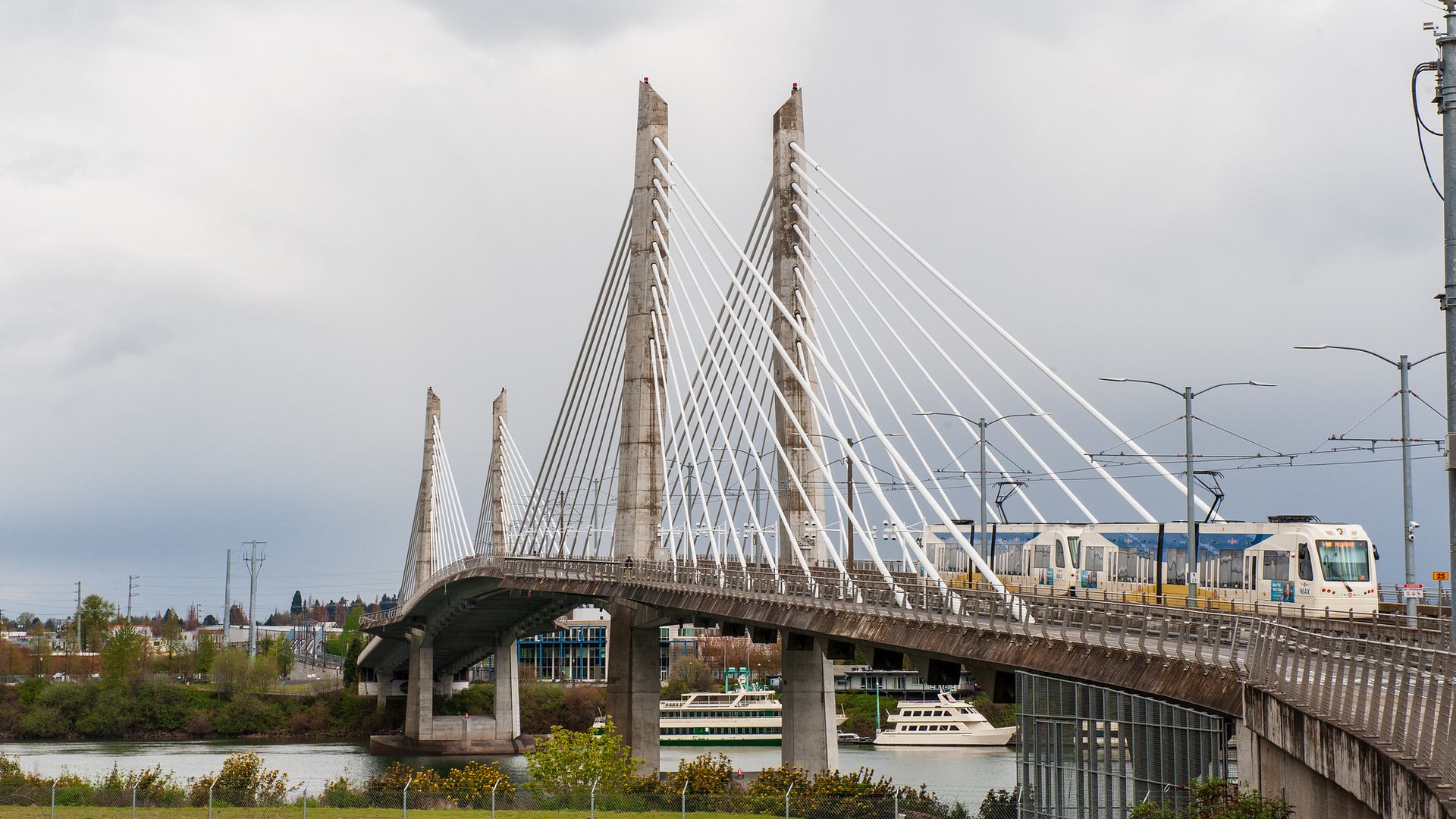 Cable-stayed bridge over water with a light rail train crossing on the right side on a cloudy day, boats docked underneath, and greenery along the water edge.
