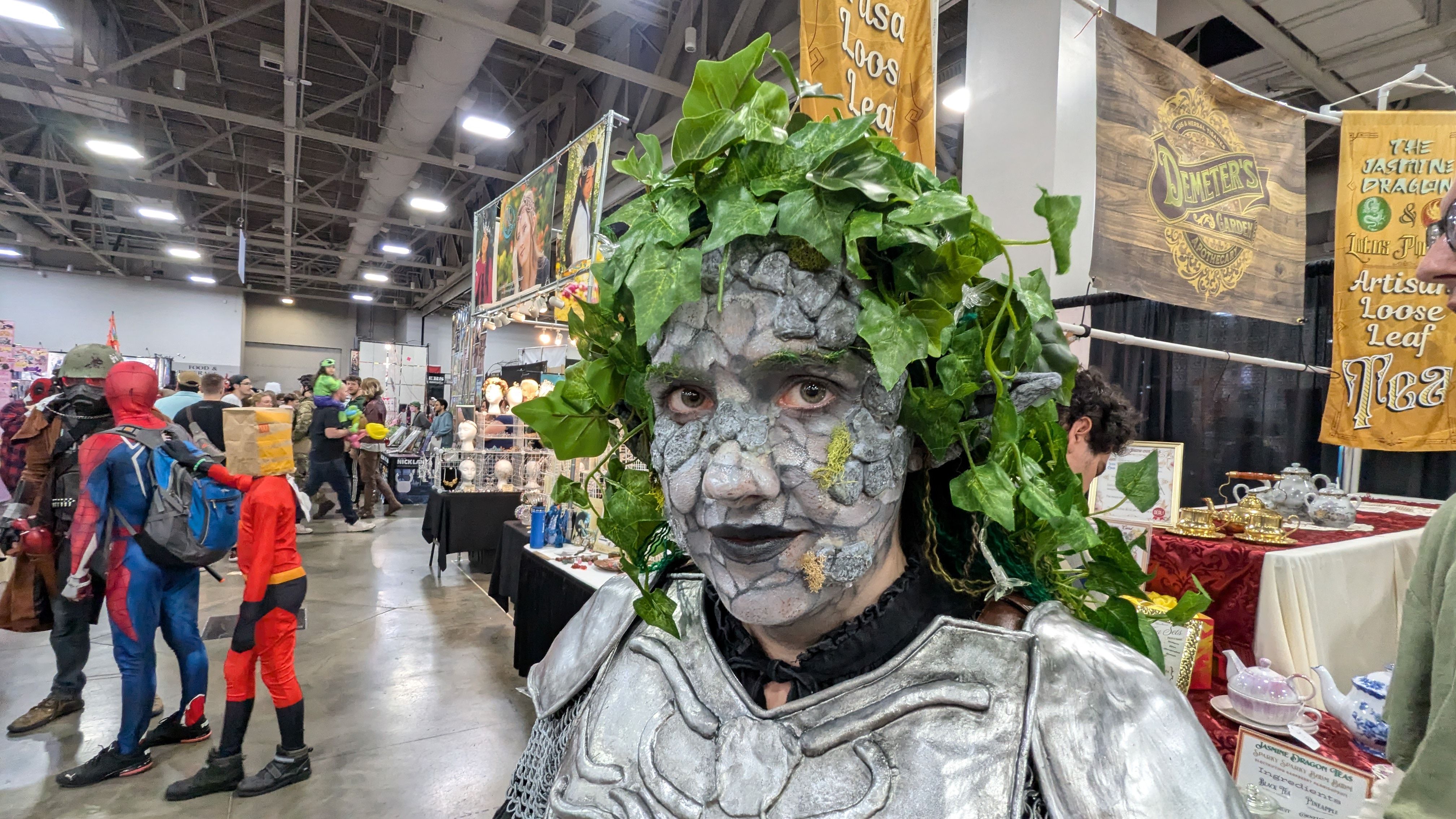 A woman with special effects makeup that causes her skin to look like stone, leaves in her hair and a coat of armor, standing in a convention center.