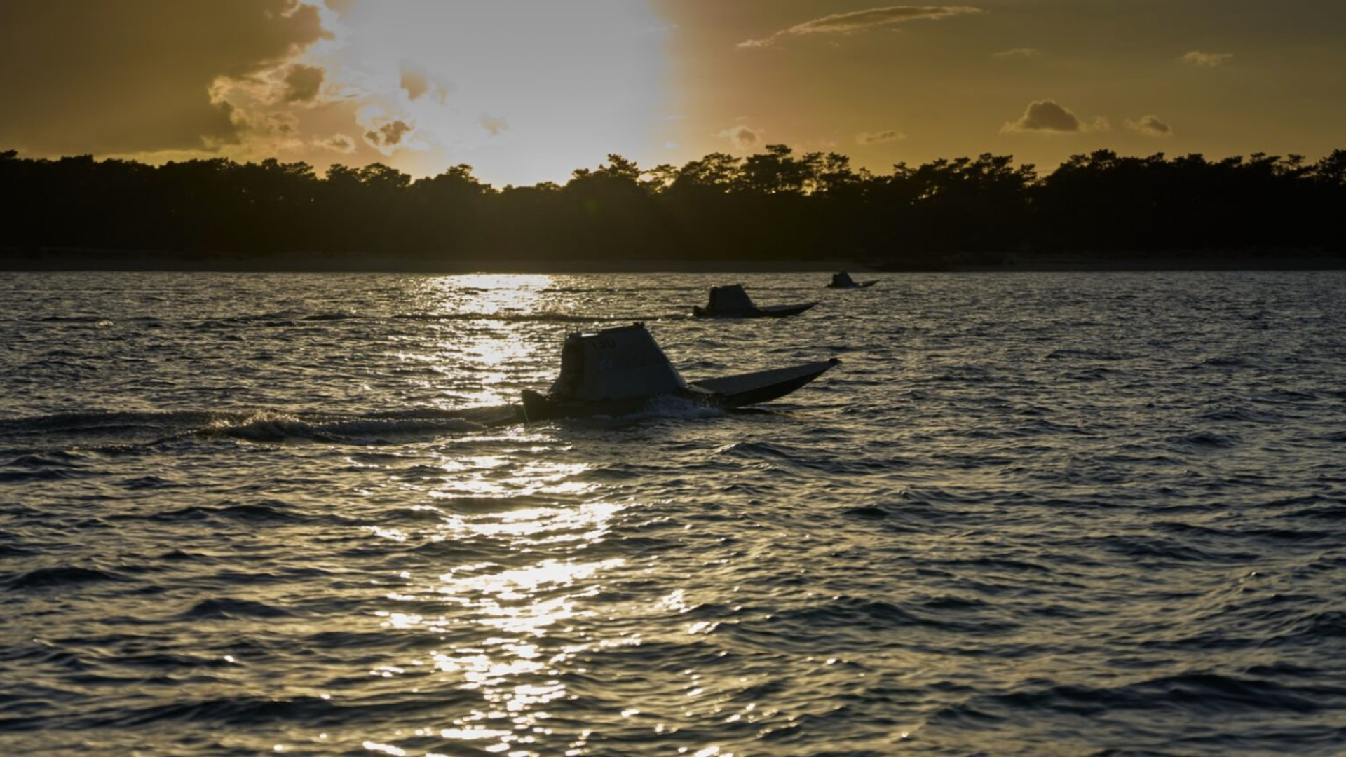 Sunset behind a silhouette of trees with three small boats moving on the water, reflecting golden light on the rippling waves.