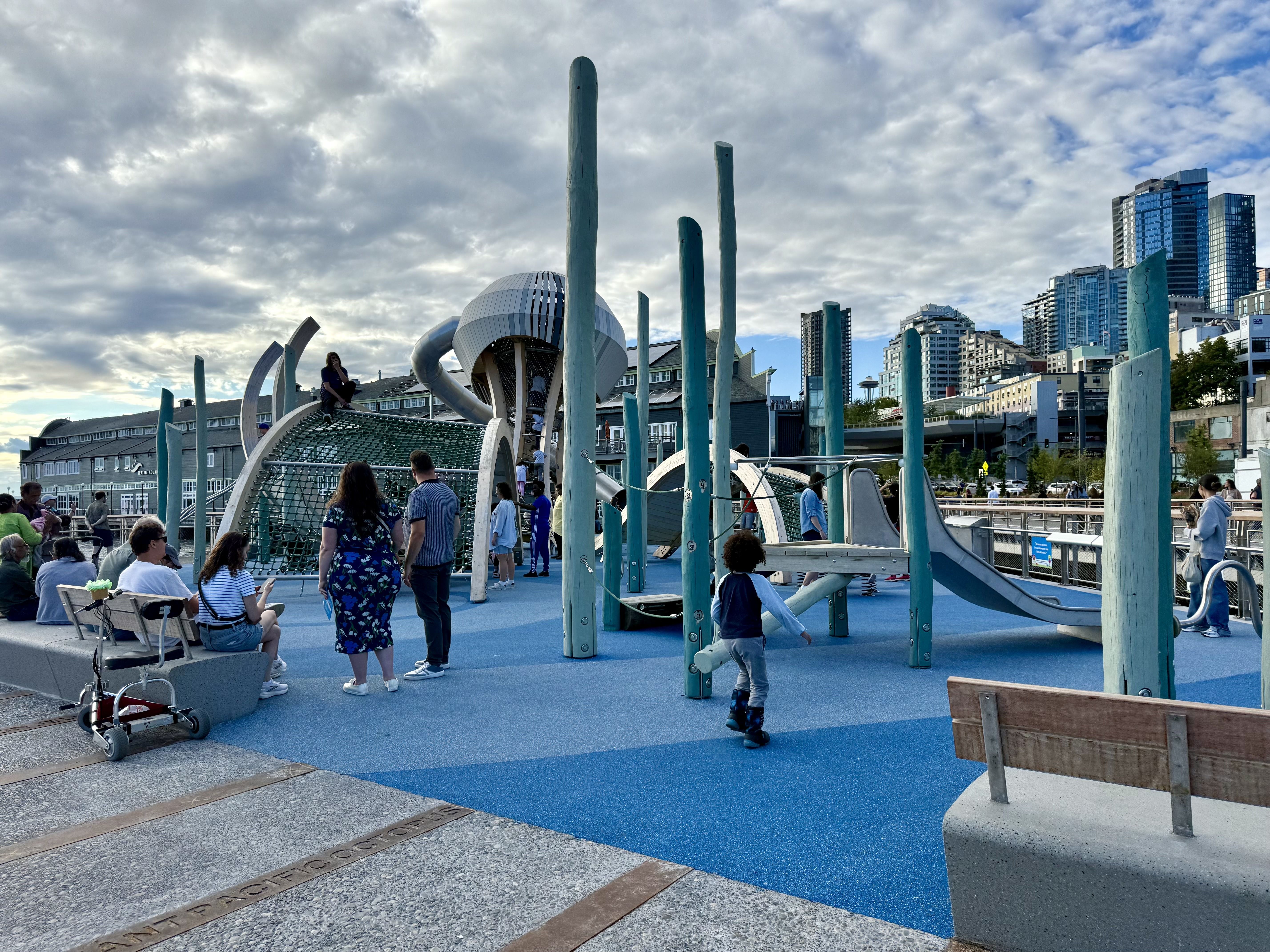 Outdoor playground with blue rubber flooring, teal vertical poles, slides, and climbing structures. Several people, including children and adults, are present under a partly cloudy sky in an urban setting.