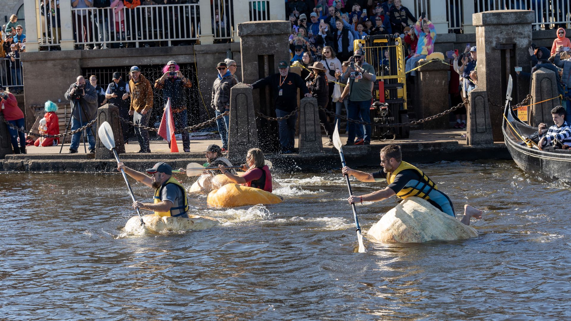Kayakers in yellow life jackets paddle through choppy river water as a crowd of spectators and photographers lines the riverbank behind railings and chains.