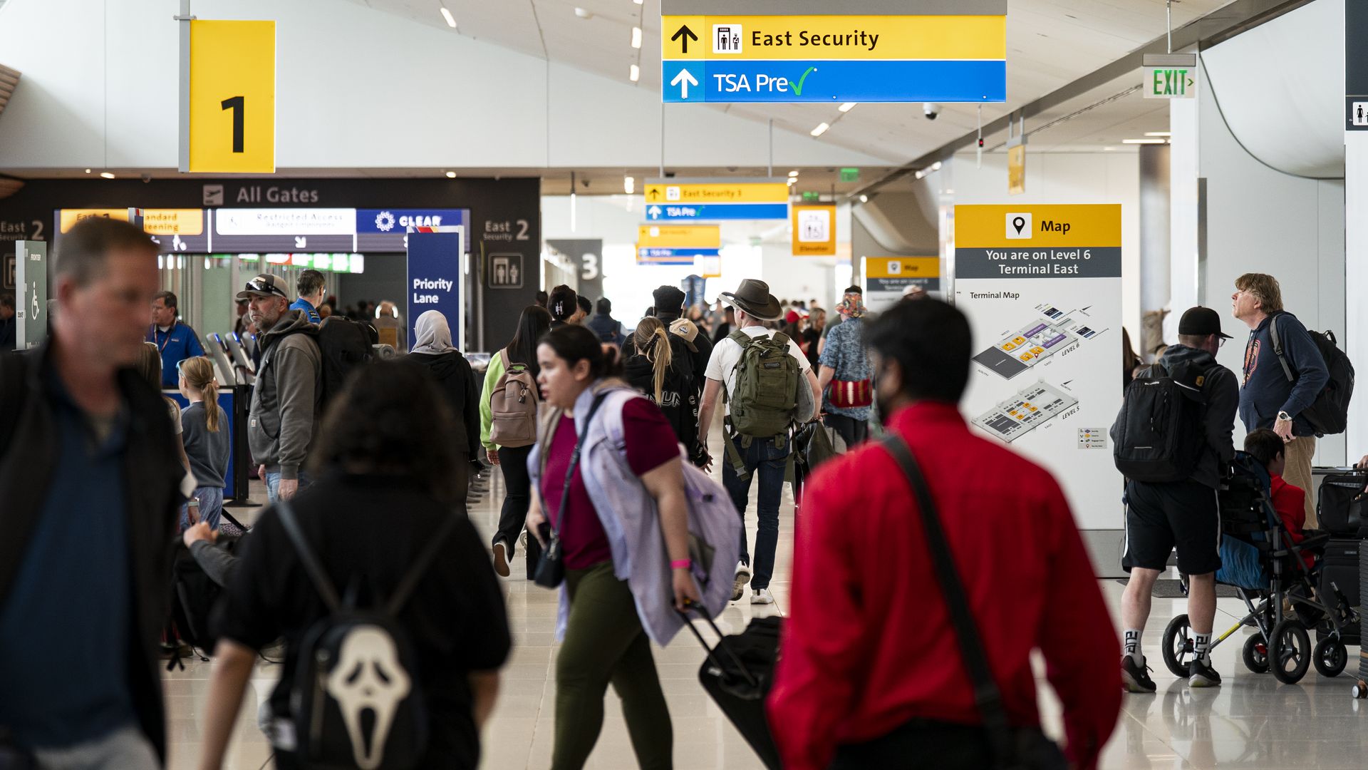 Busy airport terminal with travelers carrying backpacks and luggage. Prominent yellow and blue overhead signs show East Security and TSA Pre, large 1 gate sign left and a map sign right.