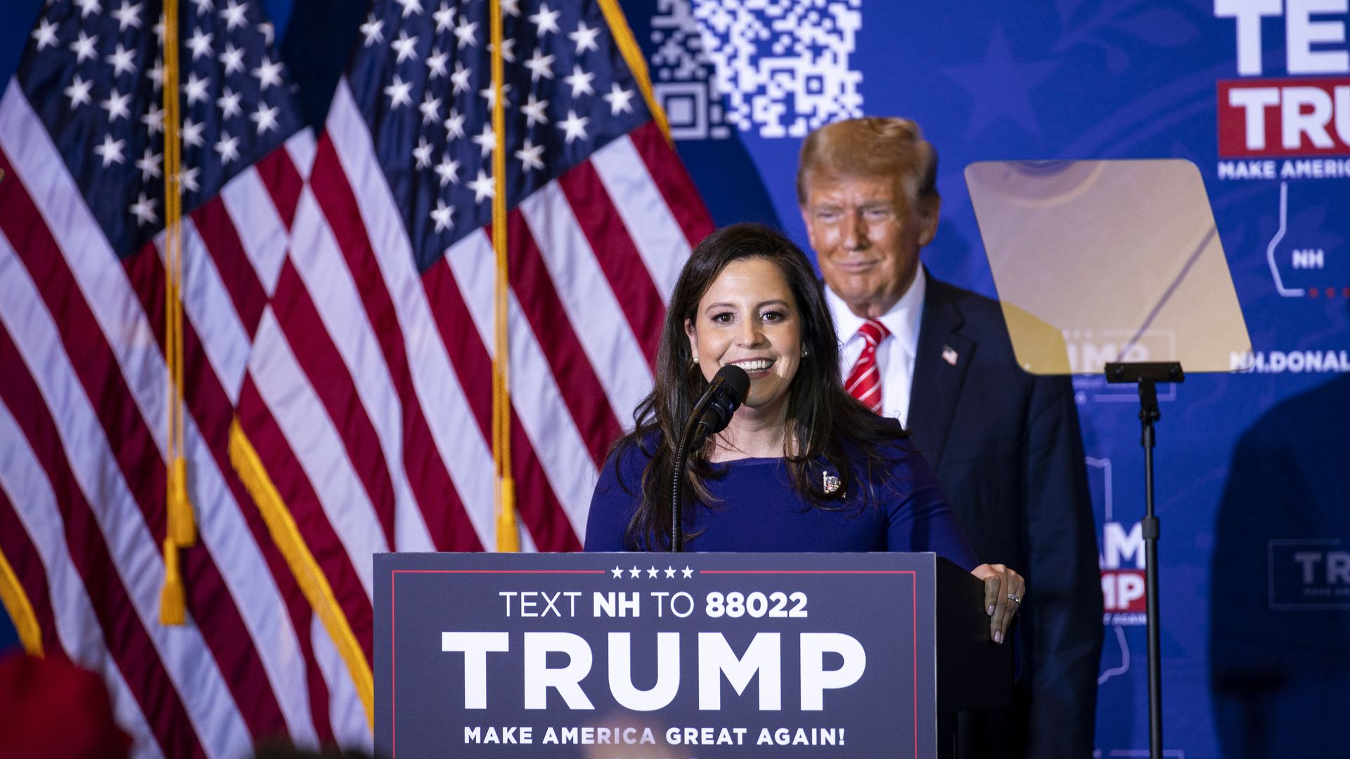 Rep. Elise Stefanik speaks behind a microphone with a big Trump sign, and Donald Trump in the background.