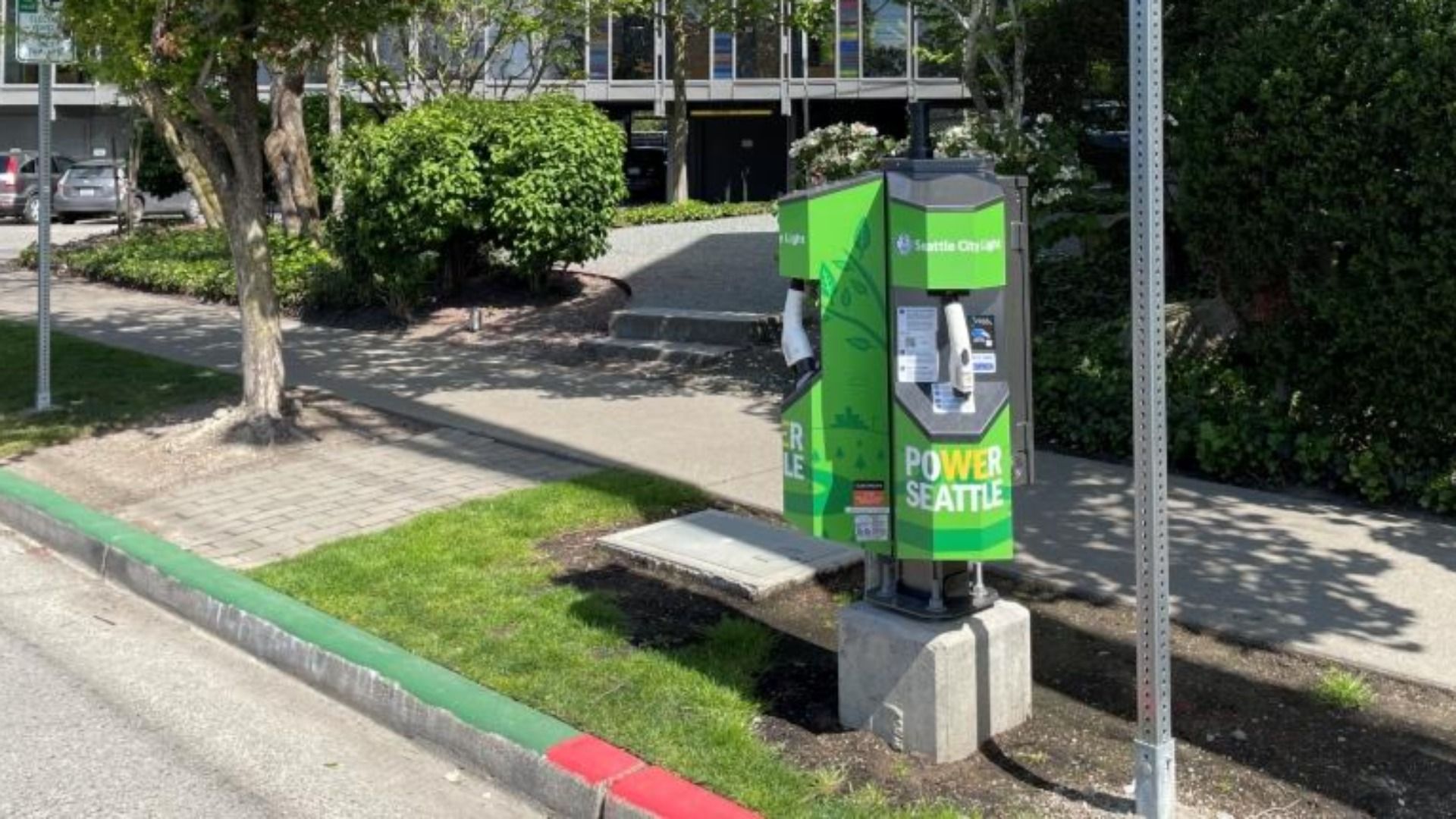 Green and gray public power kiosk on a concrete base beside a sidewalk, with Power Seattle branding, trees and shrubs nearby, and a modern building in the background; green and red curb in the foreground.