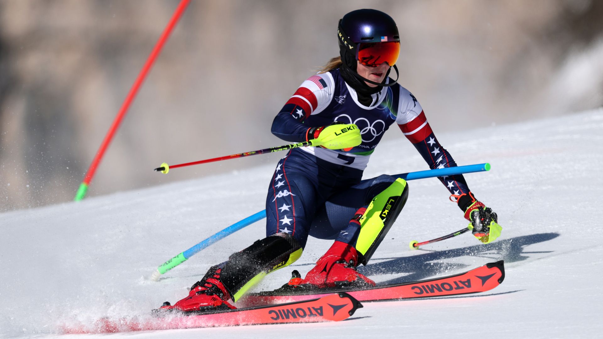 Alpine skier in USA gear and black helmet racing downhill on snowy slope, navigating blue and red slalom poles, using red Atomic skis, with Olympic rings visible on uniform.