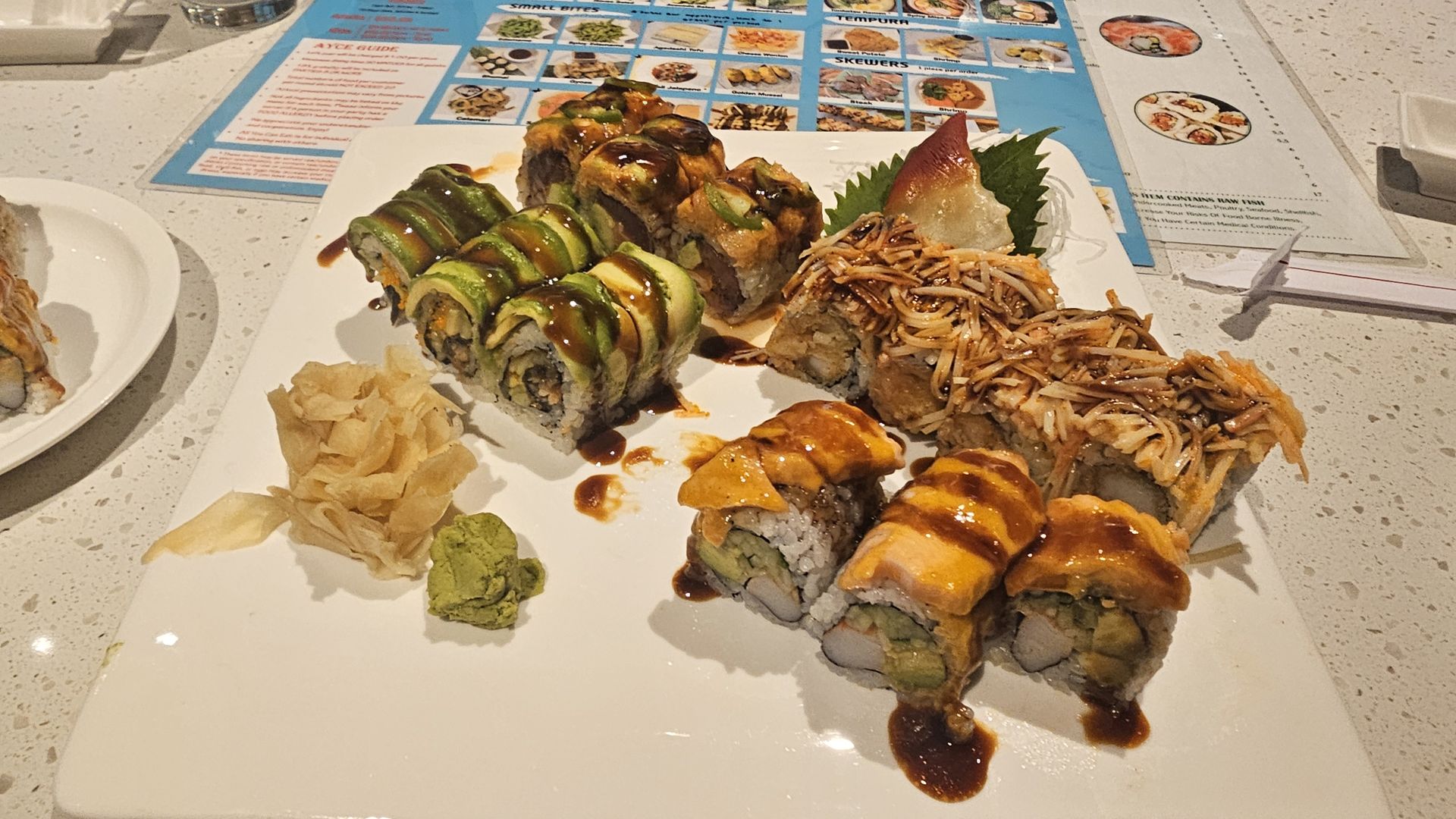Plate with various sushi rolls topped with sauces, garnished with pickled ginger and wasabi, set on a speckled white table with a menu in the background.