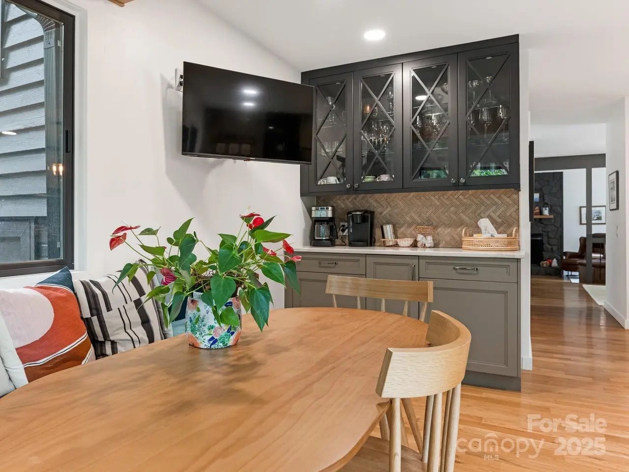 Modern dining area with wooden table and chairs, gray cabinets with glass doors, a mounted TV, colorful pillows on a bench, and a plant centerpiece under soft lighting.
