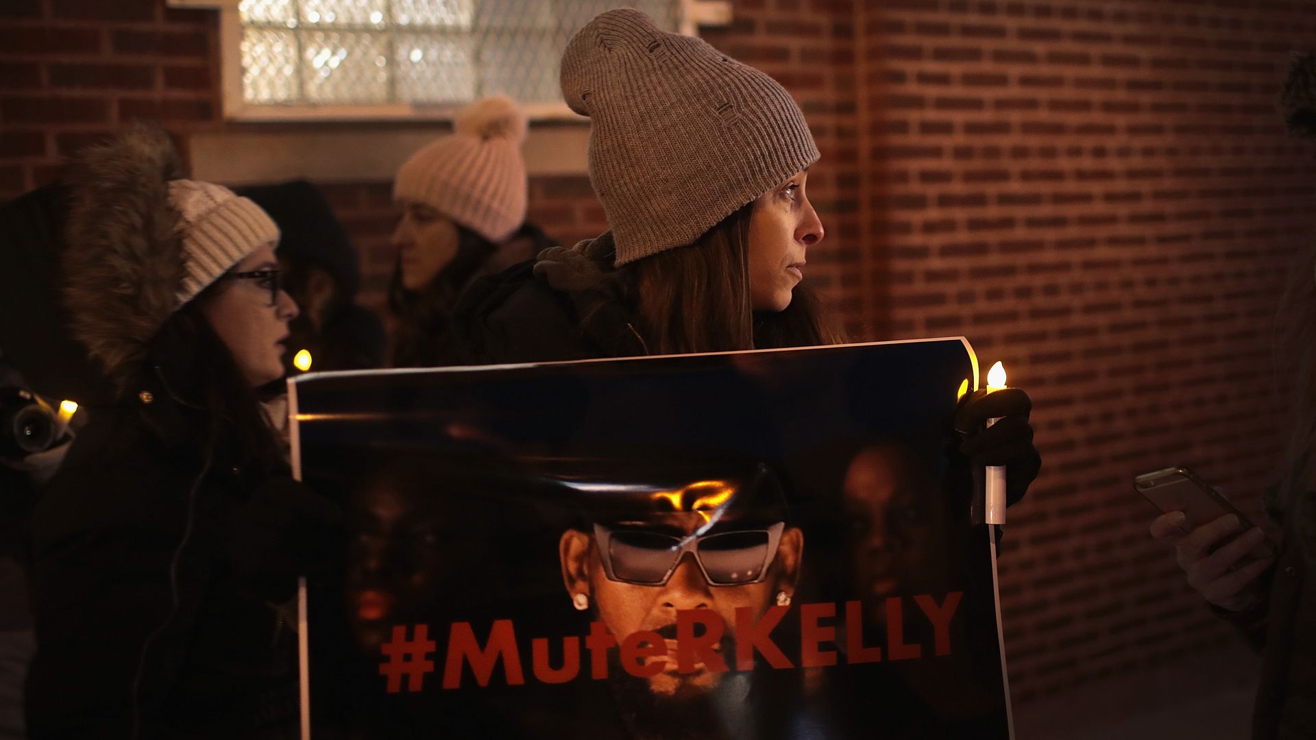 In this image, a woman in a winter coat and gloves holds a sign that says "Mute Kelly." The words on the sign are written in red over a picture of R. Kelly's face.