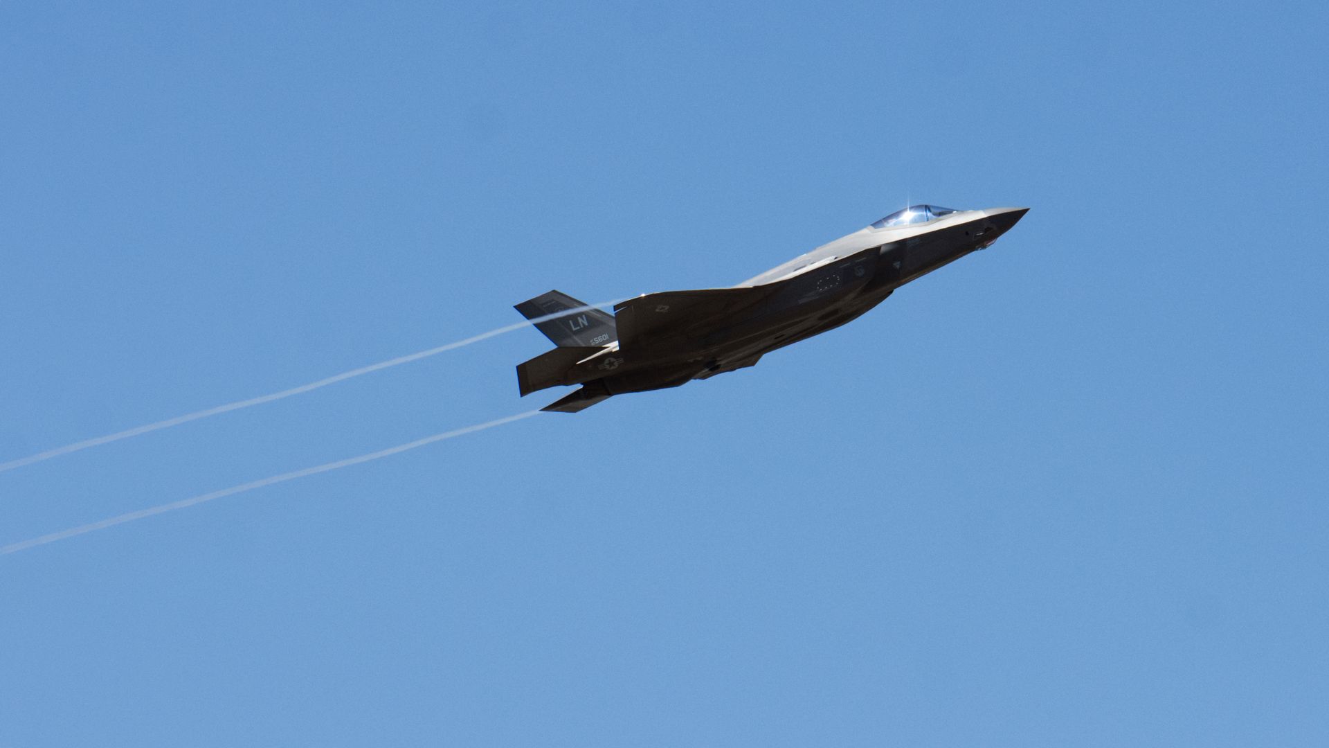 An F-35 fighter jet zips through clear blue skies. Its canopy glints in the sun.