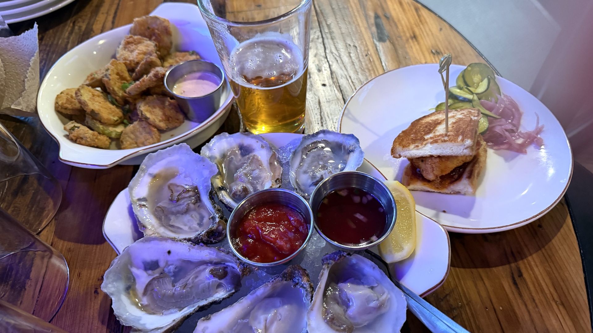 Wooden table with a feast: oysters on the half shell with lemon and sauces, a bowl of fried bites, a glass of beer, and a plate with a sandwich and pickled onions.