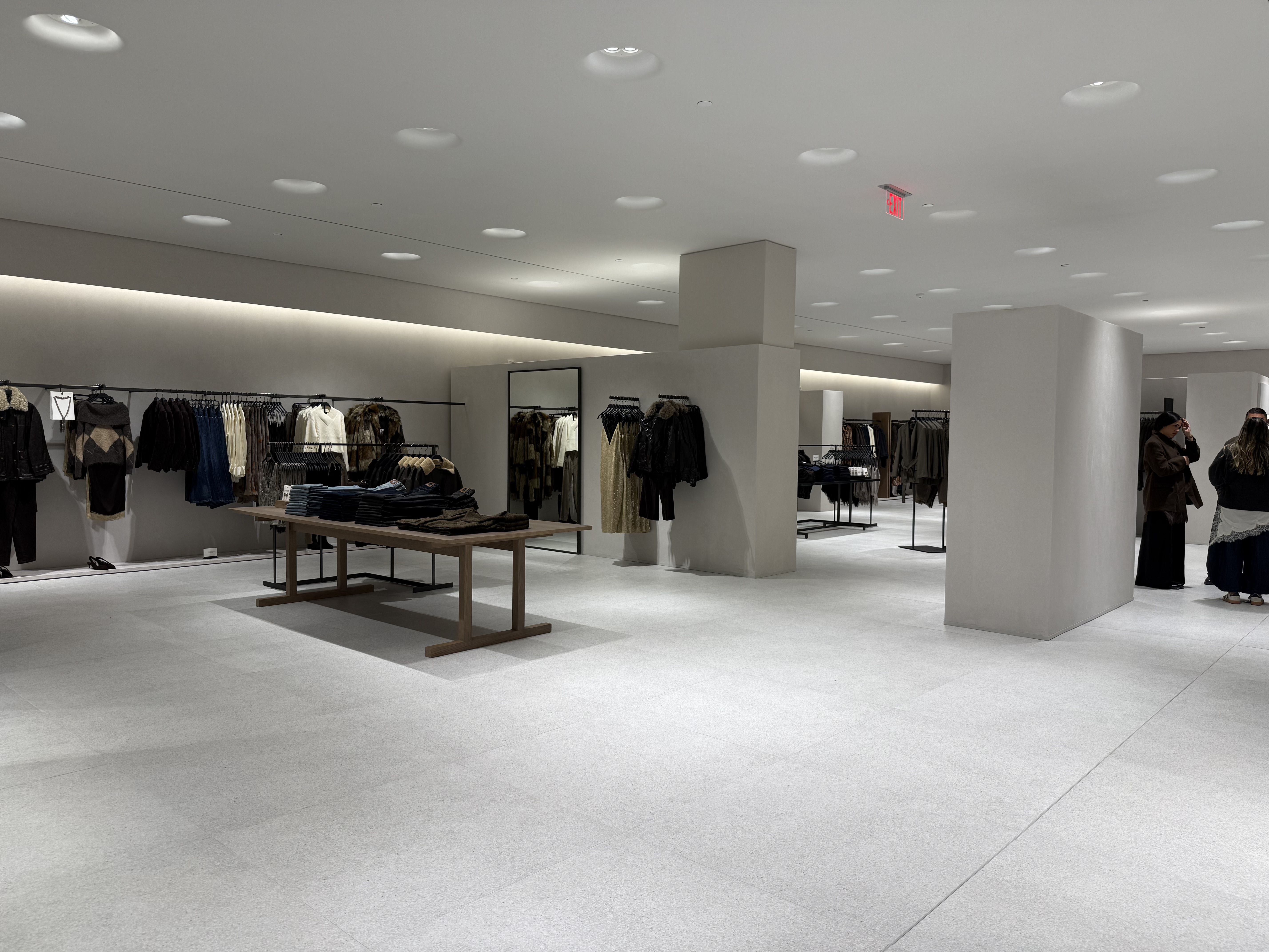 Spacious, modern clothing store with wooden display tables, racks of dark and neutral-toned clothes, light gray floors, soft ceiling lights, and three people conversing near white columns.