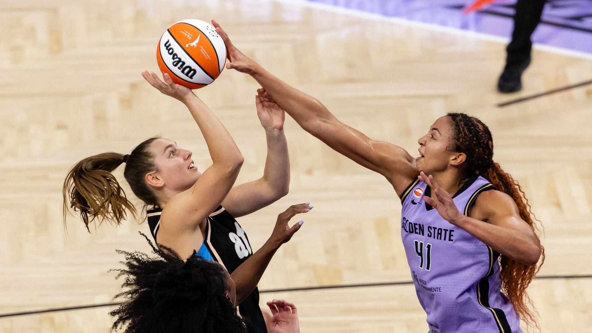 A woman basketball player wearing a violet "GOLDEN STATE" jersey blocks an attempted shot by a rival player wearing a dark black "atl" jersey.