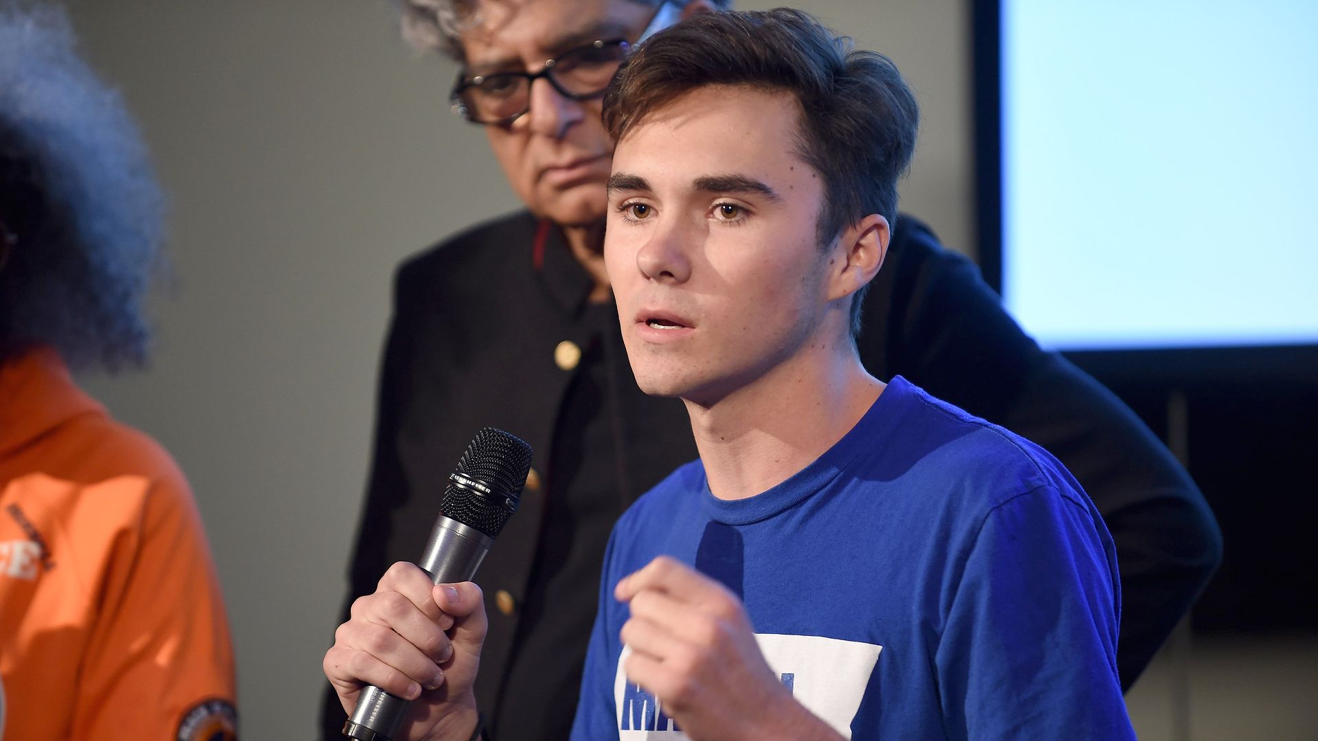 In this image, David Hogg speaks into a microphone while wearing a T shirt. 