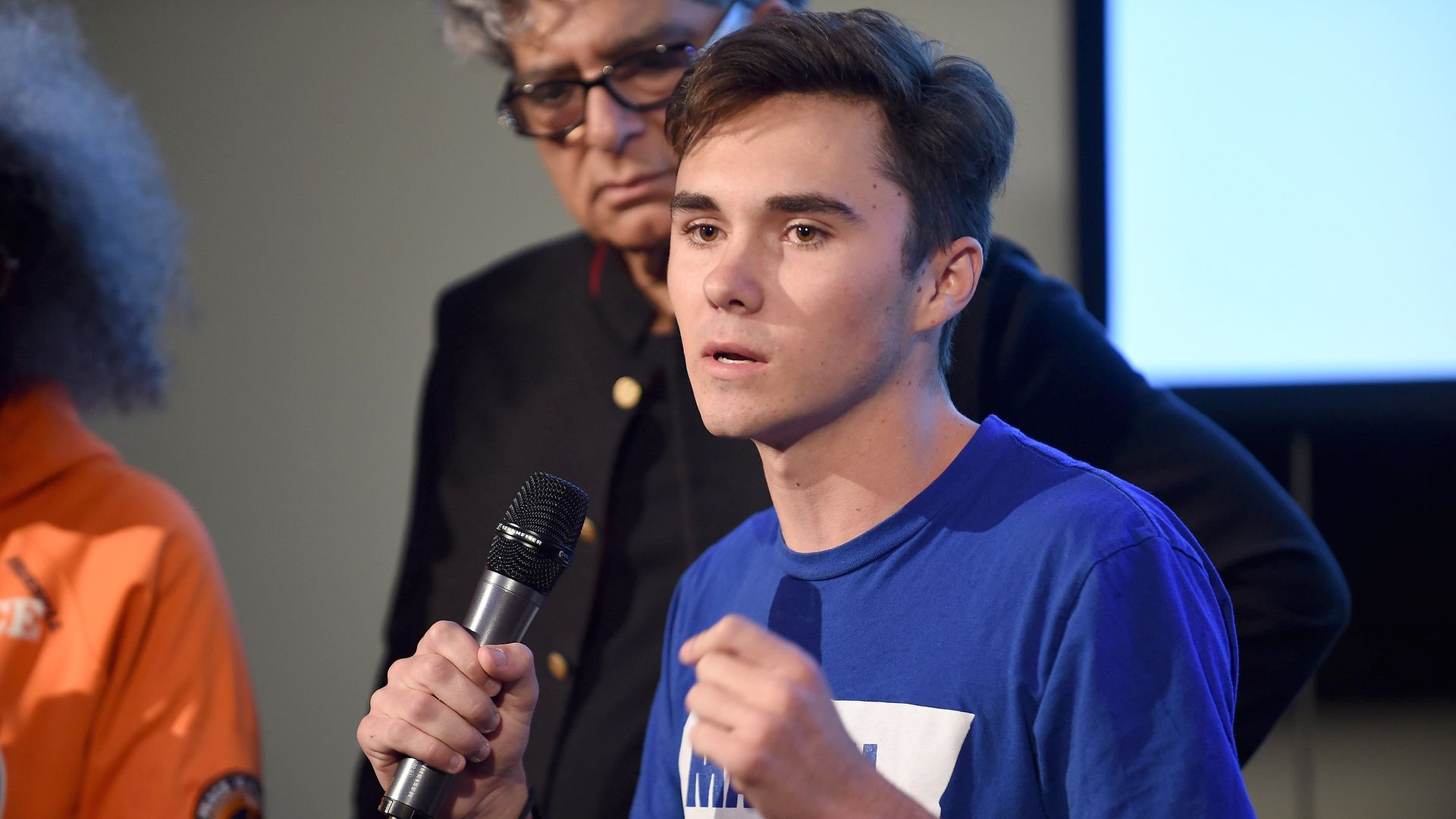 In this image, David Hogg speaks into a microphone while wearing a T shirt.
