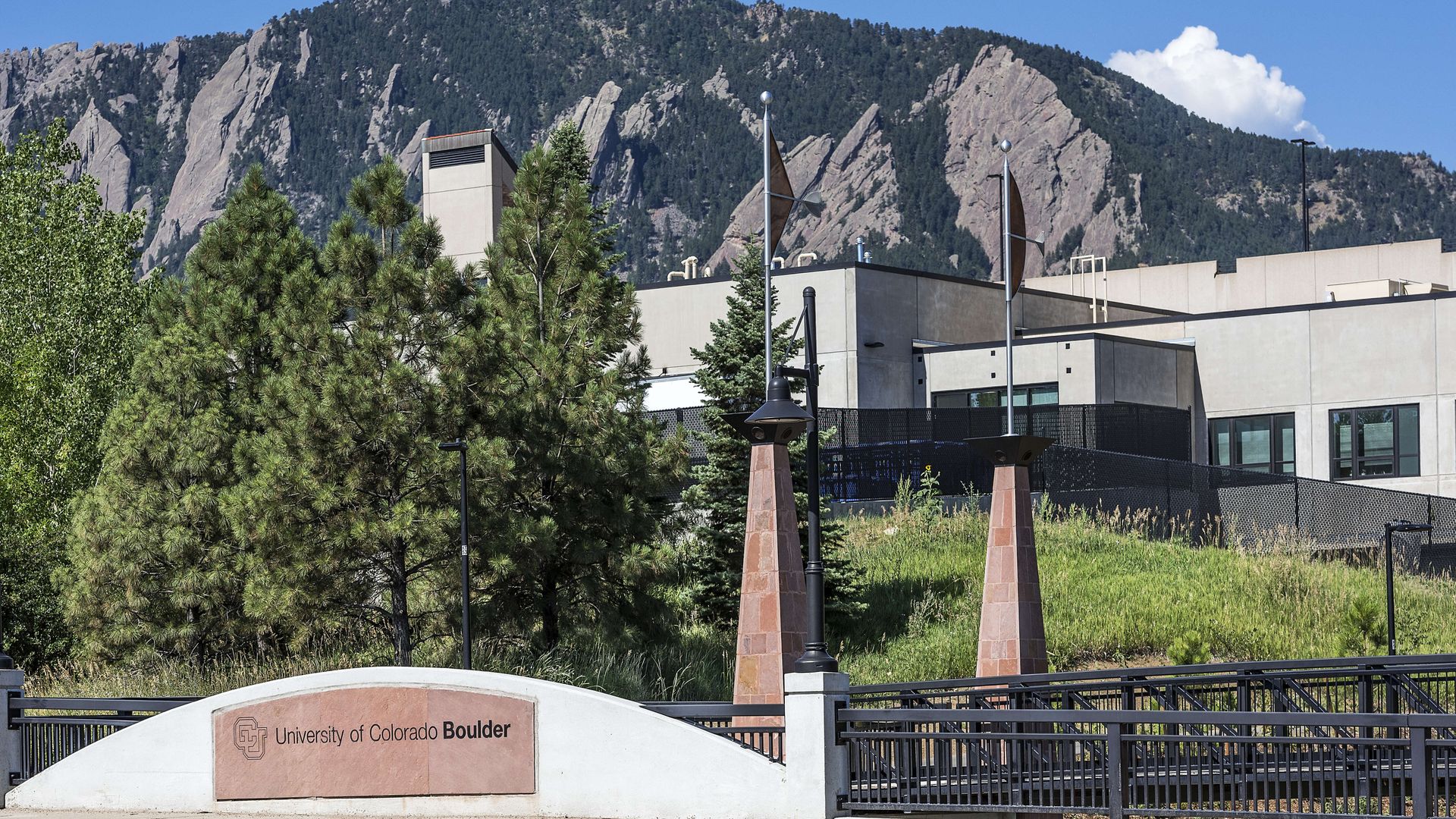 University of Colorado Boulder entrance with curved stone sign and iron fencing, green pines, beige buildings, and a rugged mountain range beneath a blue sky.