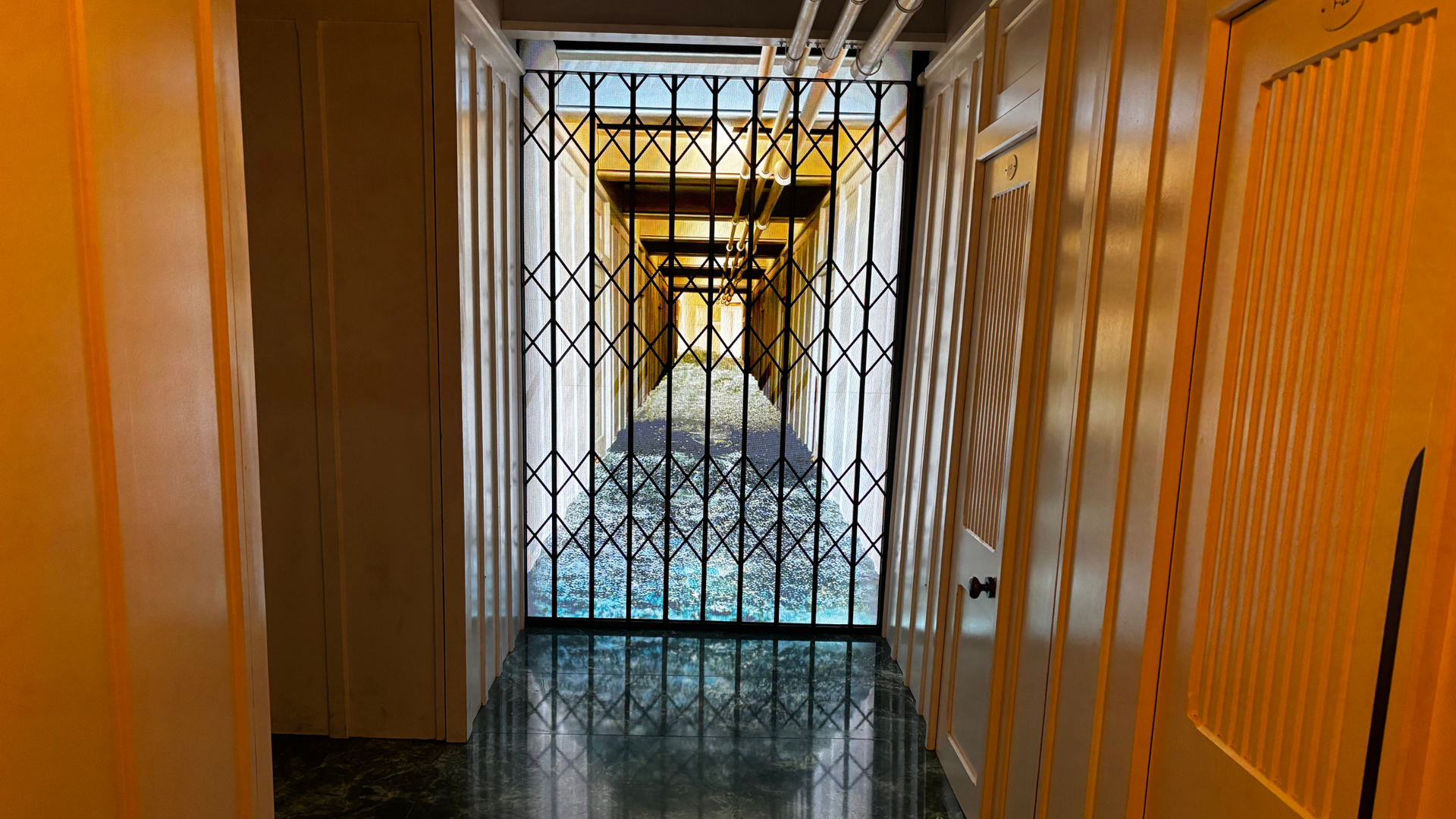 Narrow hallway with orange walls, a black diamond-pattern metal gate at the far end, overhead pipes, and a glossy dark marble floor reflecting light.