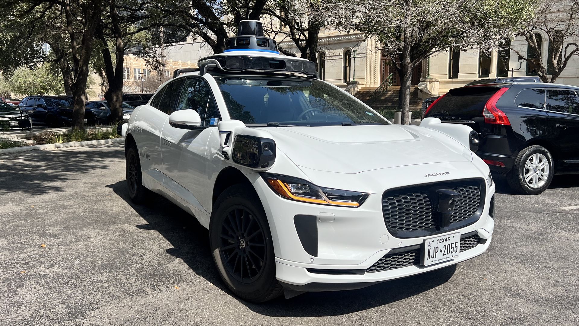 White Jaguar electric car equipped with sensors and cameras for autonomous driving, parked outdoors under trees with other cars nearby and a building in the background.