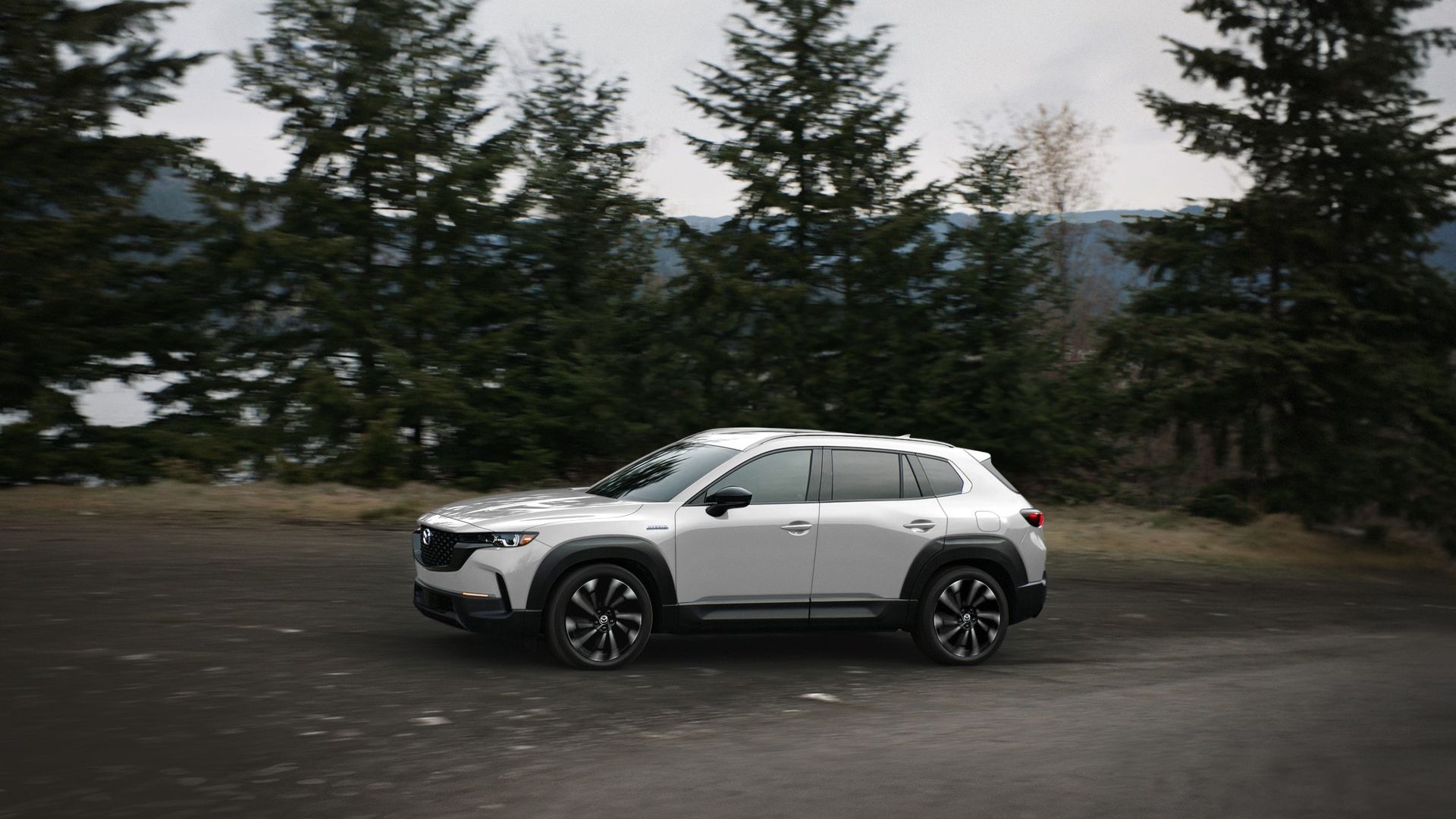 Image of a white Mazda CX-50 crossover utility parking near some pine trees on the shore of a lake. 