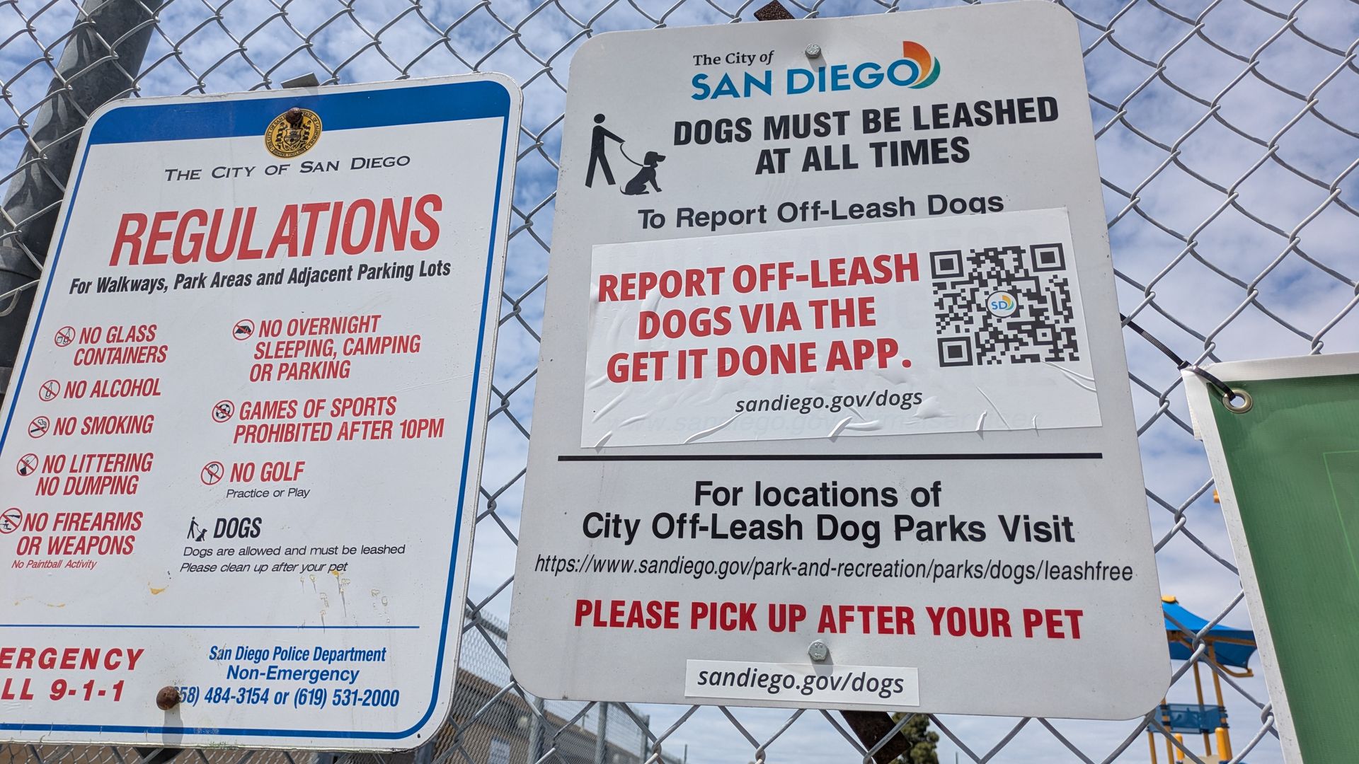 Signs posted on a chain-link fence: a San Diego regulations sign with large red REGULATIONS and another sign stating DOGS MUST BE LEASHED AT ALL TIMES with the city logo; blue sky behind.