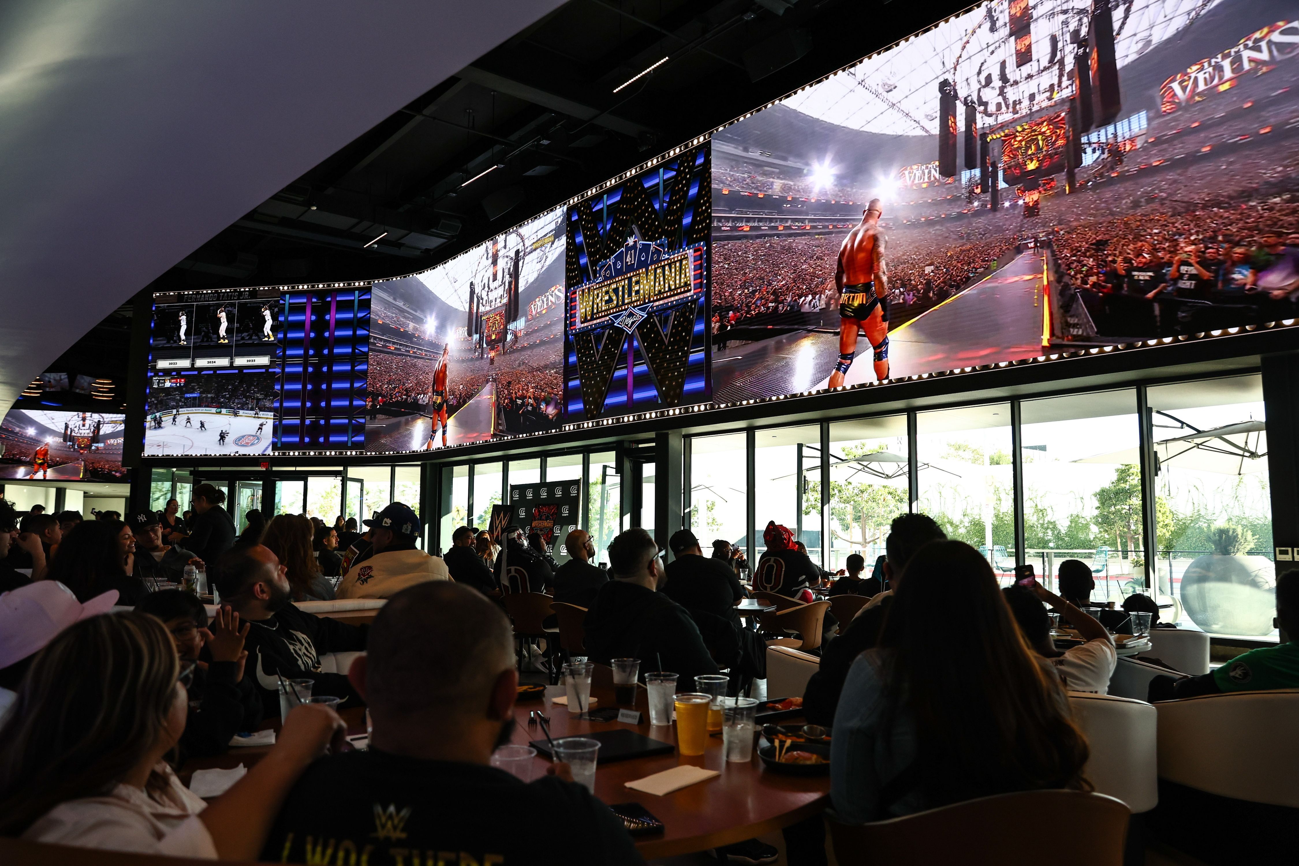 People seated indoors watching a large curved screen displaying a WWE WrestleMania event with a wrestler walking toward a huge, packed stadium crowd.