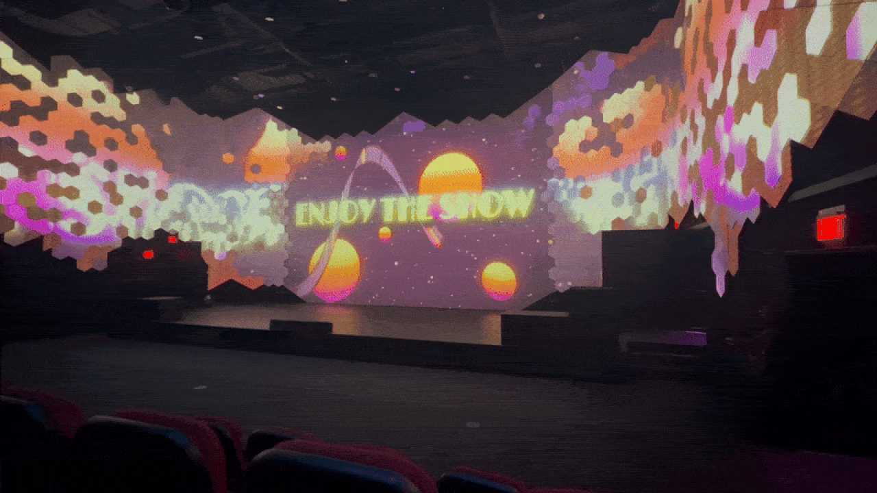 Dark theater with colorful pixelated projections on walls and stage, featuring orange and purple tones with suns and the words "ENJOY THE SHOW" in bright yellow.