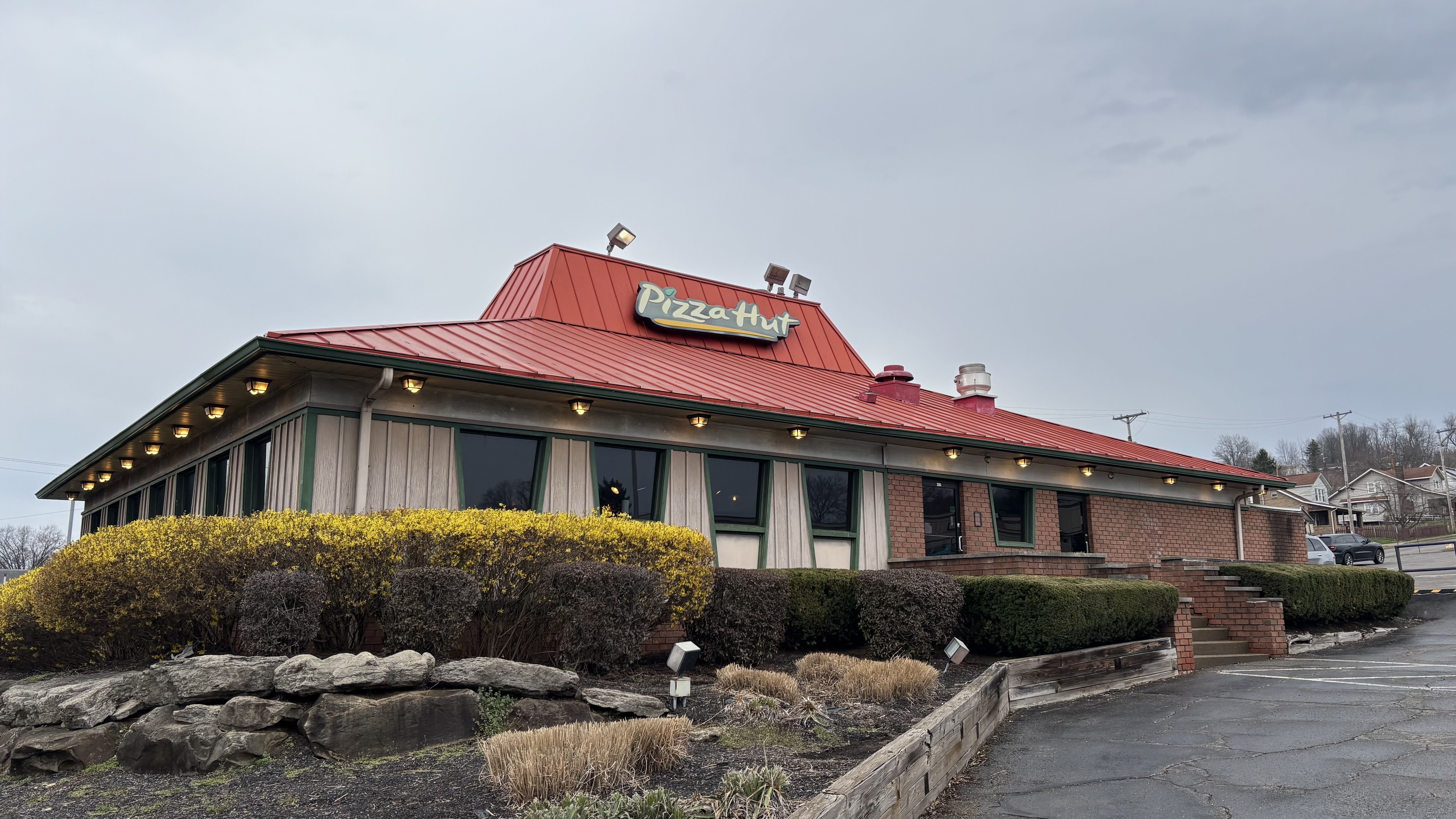 A Pizza Hut restaurant with a bright red sloped roof and green-trim exterior, large windows, and a yellow shrub border in the foreground; gray cloudy sky overhead, with a few cars in the parking lot.