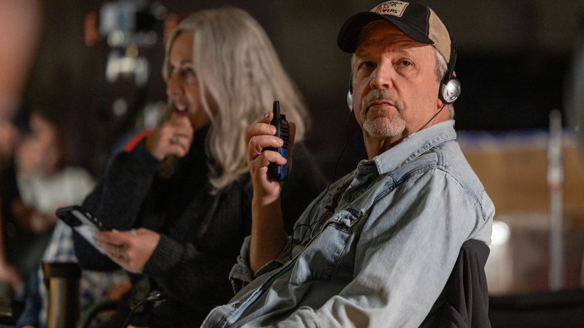 Two people in a dim setting: a serious man in a beige cap, denim jacket, and headset in the foreground holding a small radio; a smiling silver-haired woman in the background.