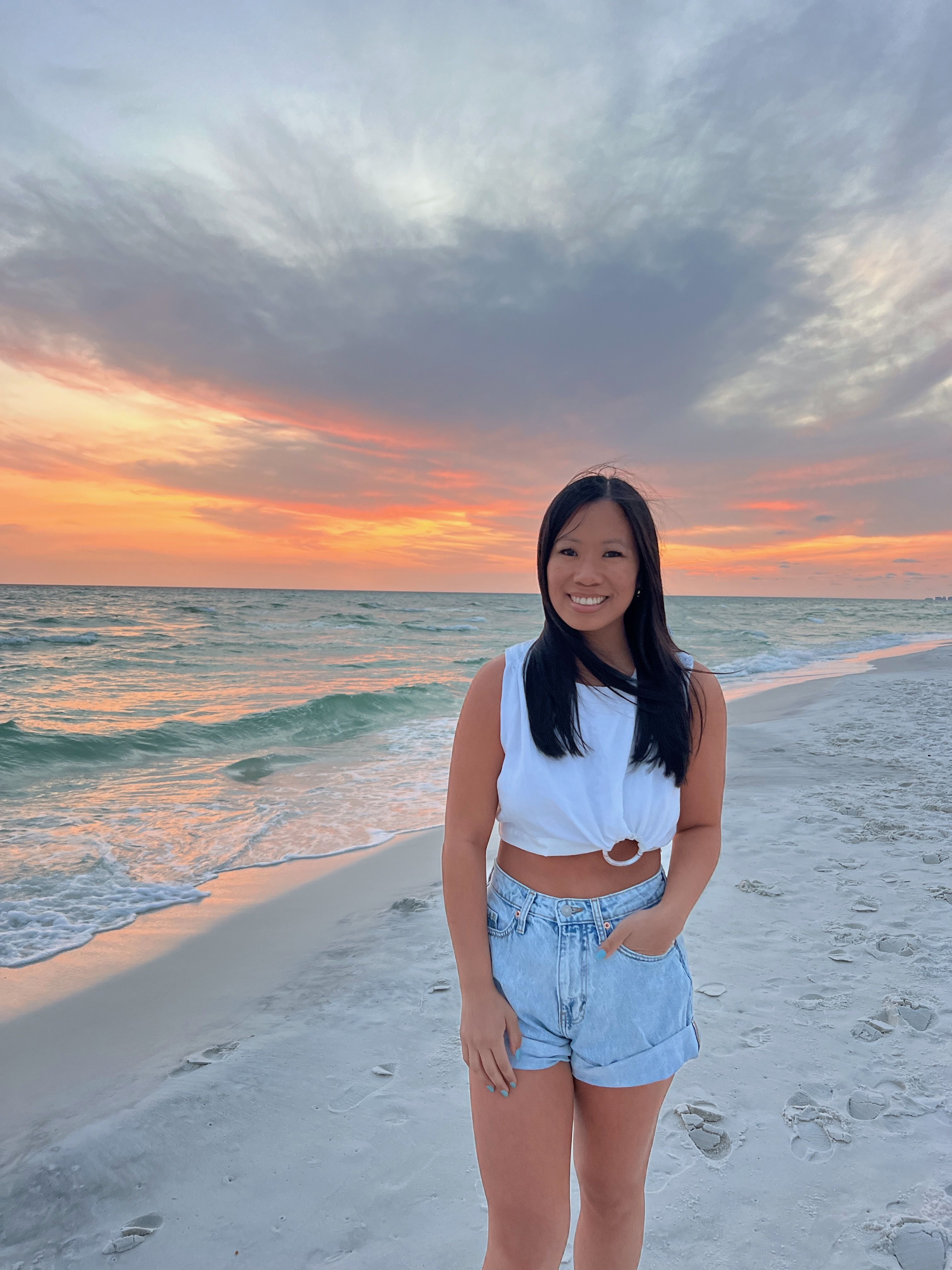 Smiling woman stands on a sandy beach at sunset, wearing a white sleeveless top and light denim shorts, with the ocean waves and a warm orange-pink sky behind her.