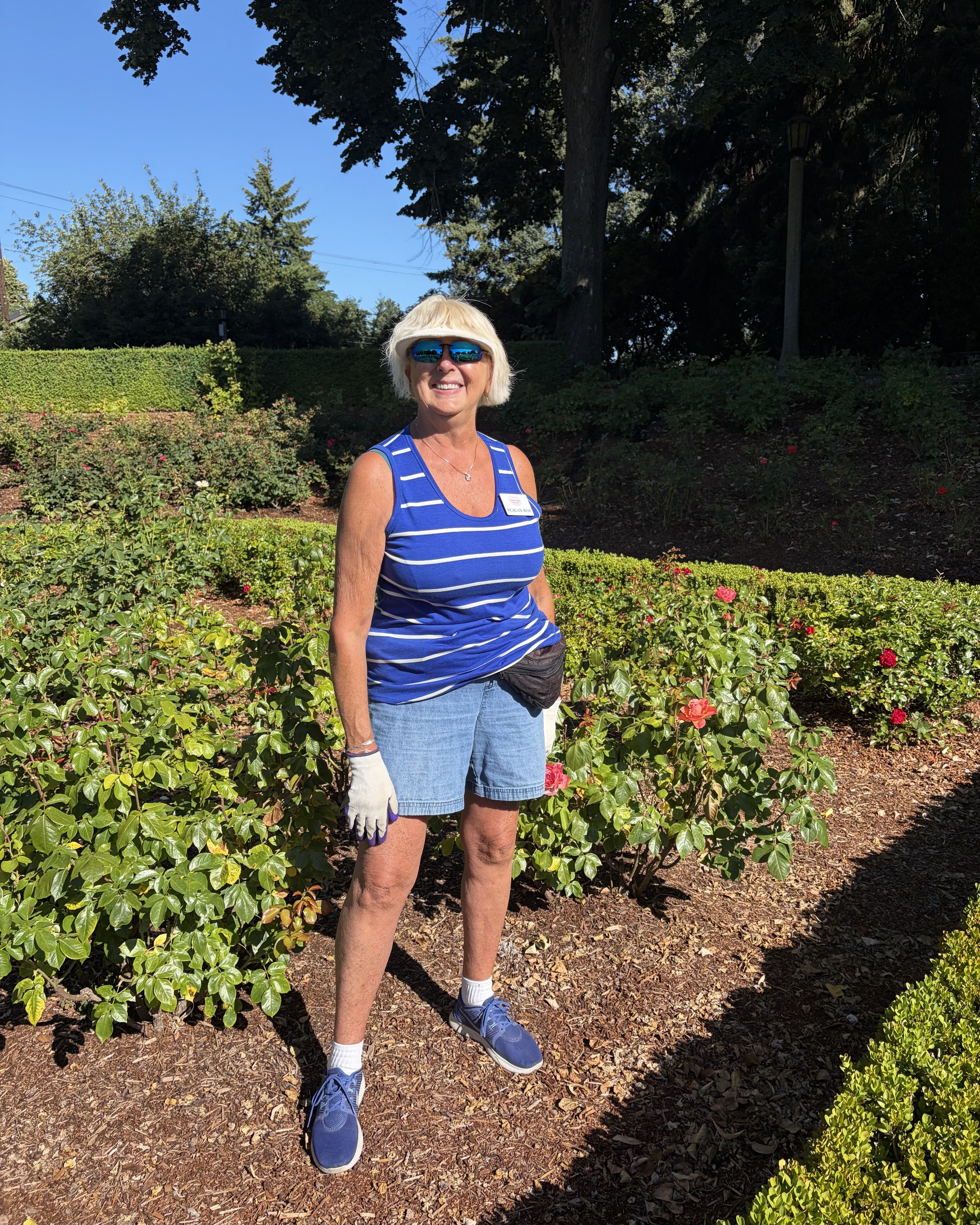 Smiling woman in a blue and white striped tank top, denim shorts, sunglasses, purple gloves, and a small waist pouch stands among rose bushes in a sunny garden.