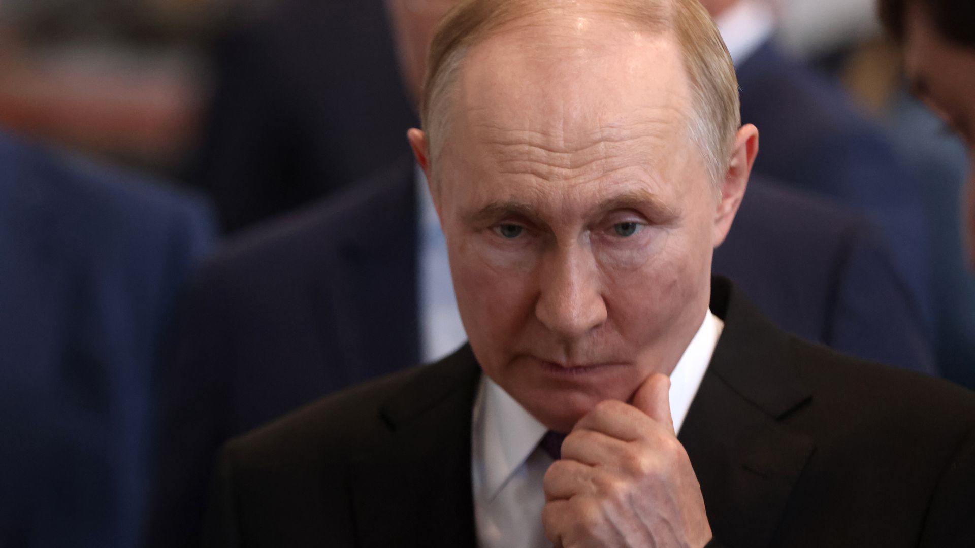 Middle-aged man with light skin and receding hair in a black suit and purple tie, holding his chin thoughtfully in an indoor setting with blurred people in the background.
