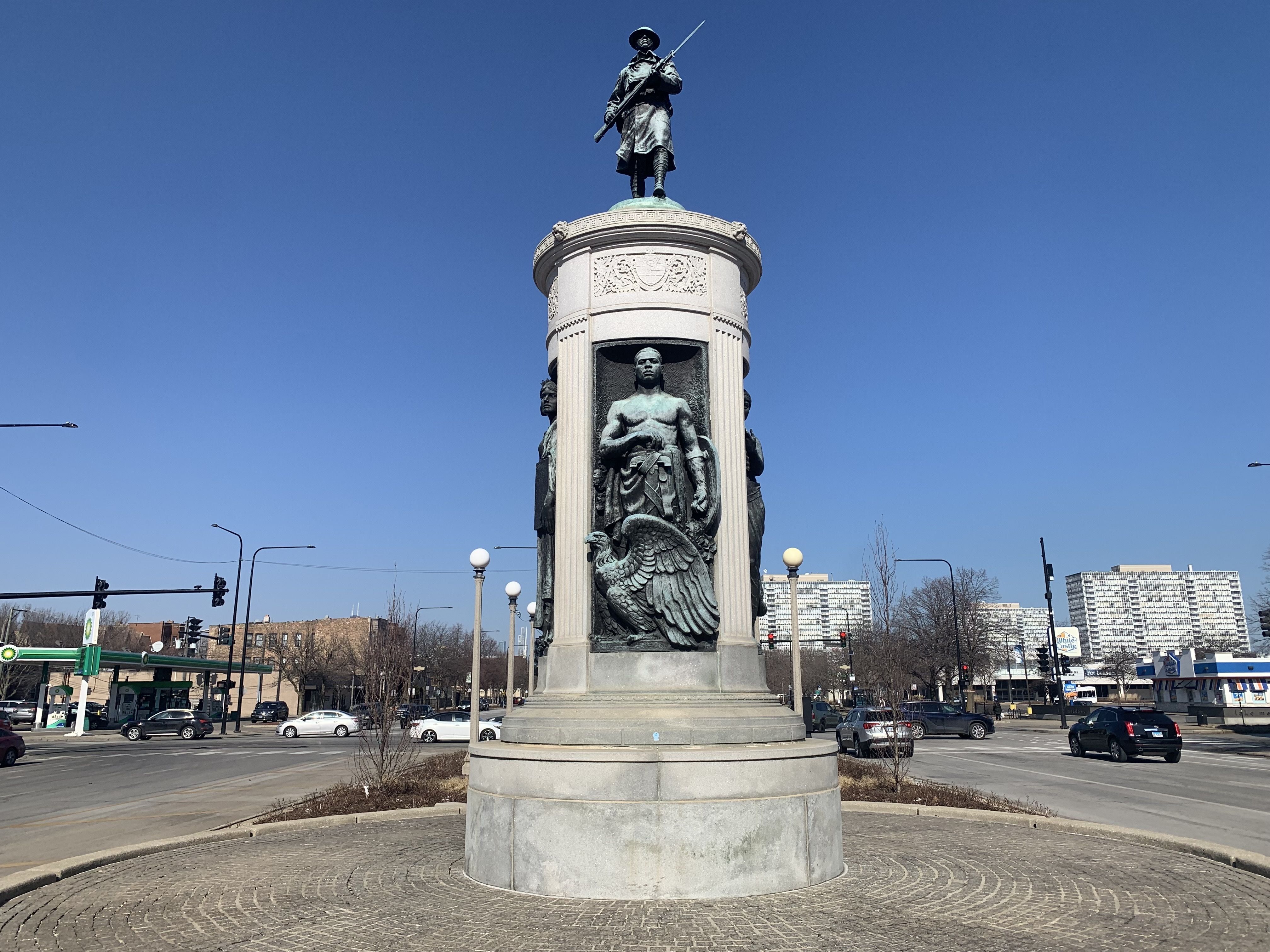 A tall monument with bronze sculptures of soldiers in a traffic circle, under a clear blue sky, surrounded by cars, streetlights, and buildings in the background.
