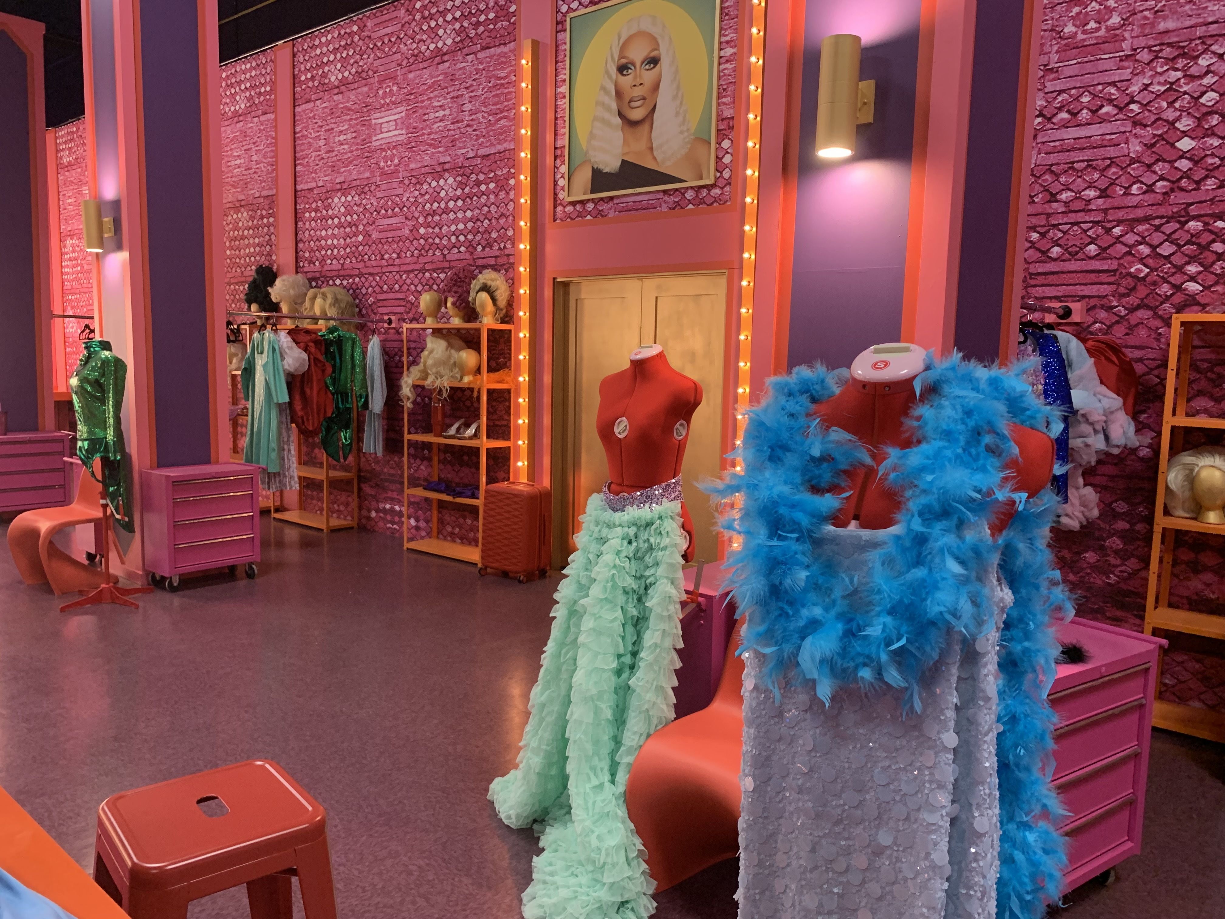 Colorful dressing room with pink and purple walls, mannequins dressed in a ruffled mint green skirt and a sequined outfit with a blue feather boa, wigs on shelves, and bright lights.