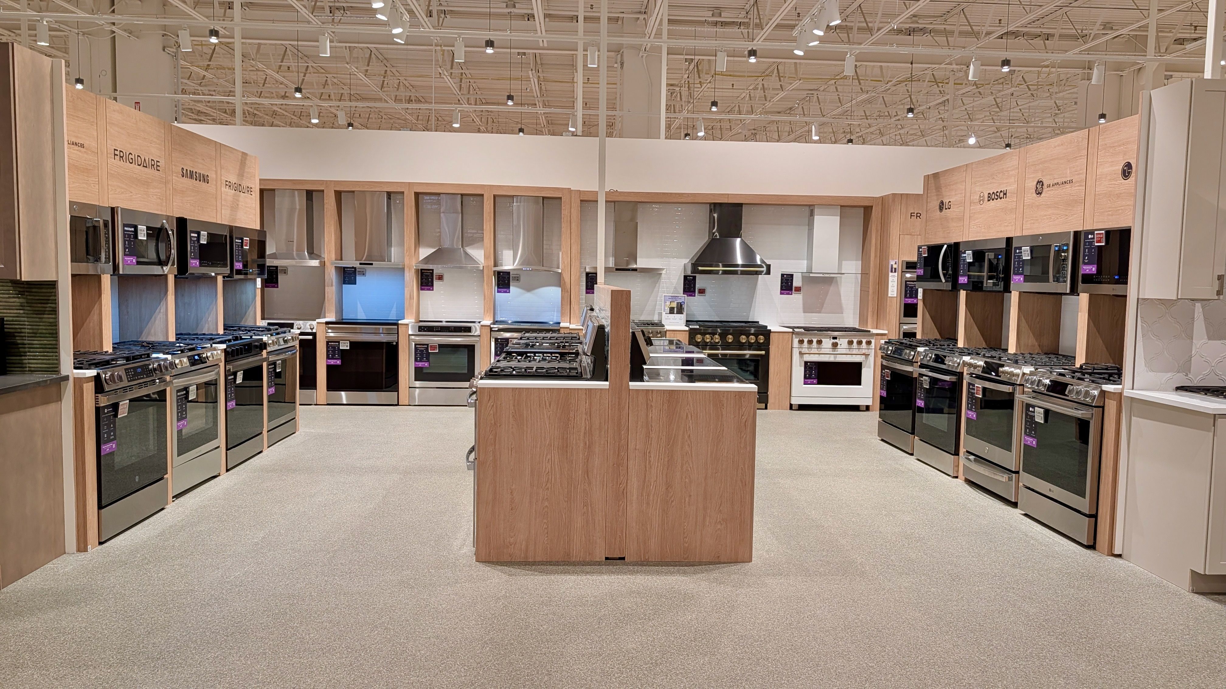 Bright appliance showroom with wooden displays along both walls, showcasing stainless steel ovens, microwaves, and range hoods; a central wooden island.