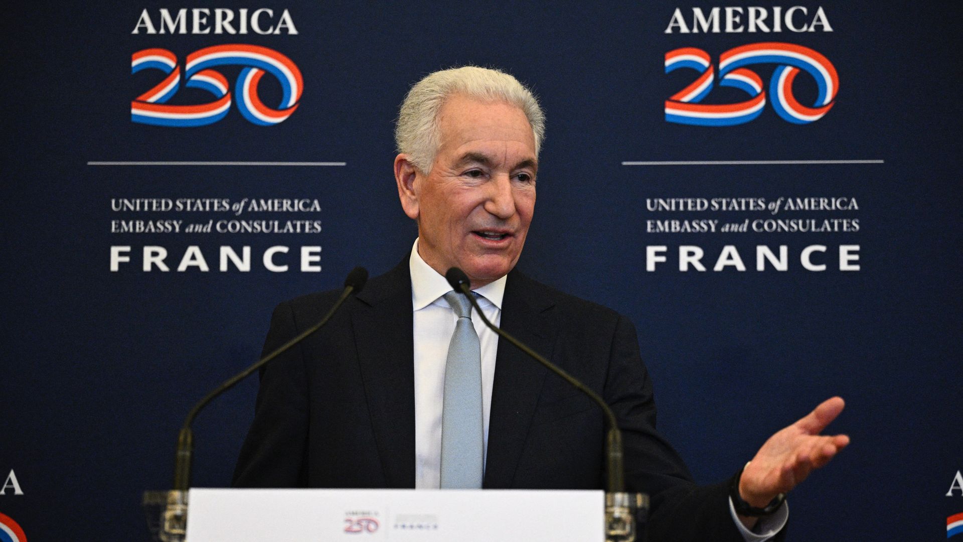 A man with white hair — wearing a black suit, a white collared shirt and a blue tie — gestures as he speaks into two microphones on a lectern with the words "250: UNITED STATES of AMERICA EMBASSY and CONSULATES FRANCE" on the backdrop behind him.
