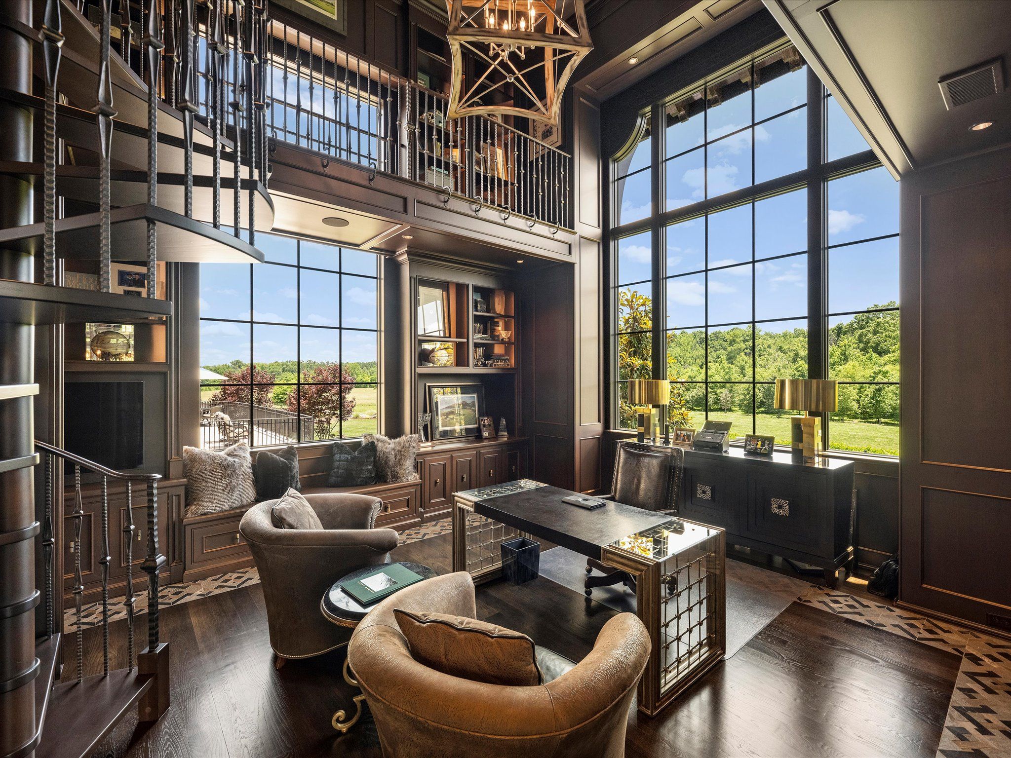 Elegant home office with dark wood walls, large grid windows showing green landscape, leather armchairs, modern desk with mirrored sides, built-in shelves, and spiral staircase.