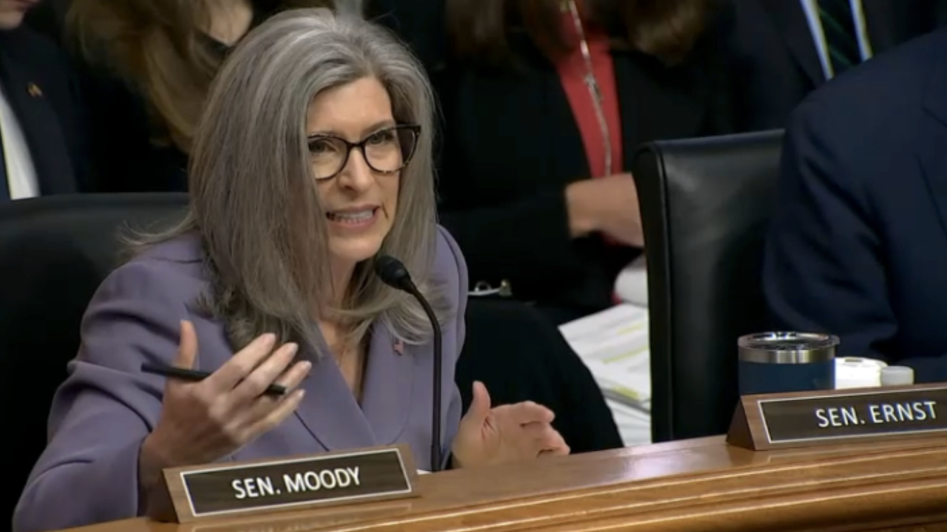 U.S. Sen. Joni Ernst with glasses in a lavender blazer speaks into a microphone at a wooden desk; visible nameplate reads SEN. MOODY and another sign SEN. ERNST nearby.