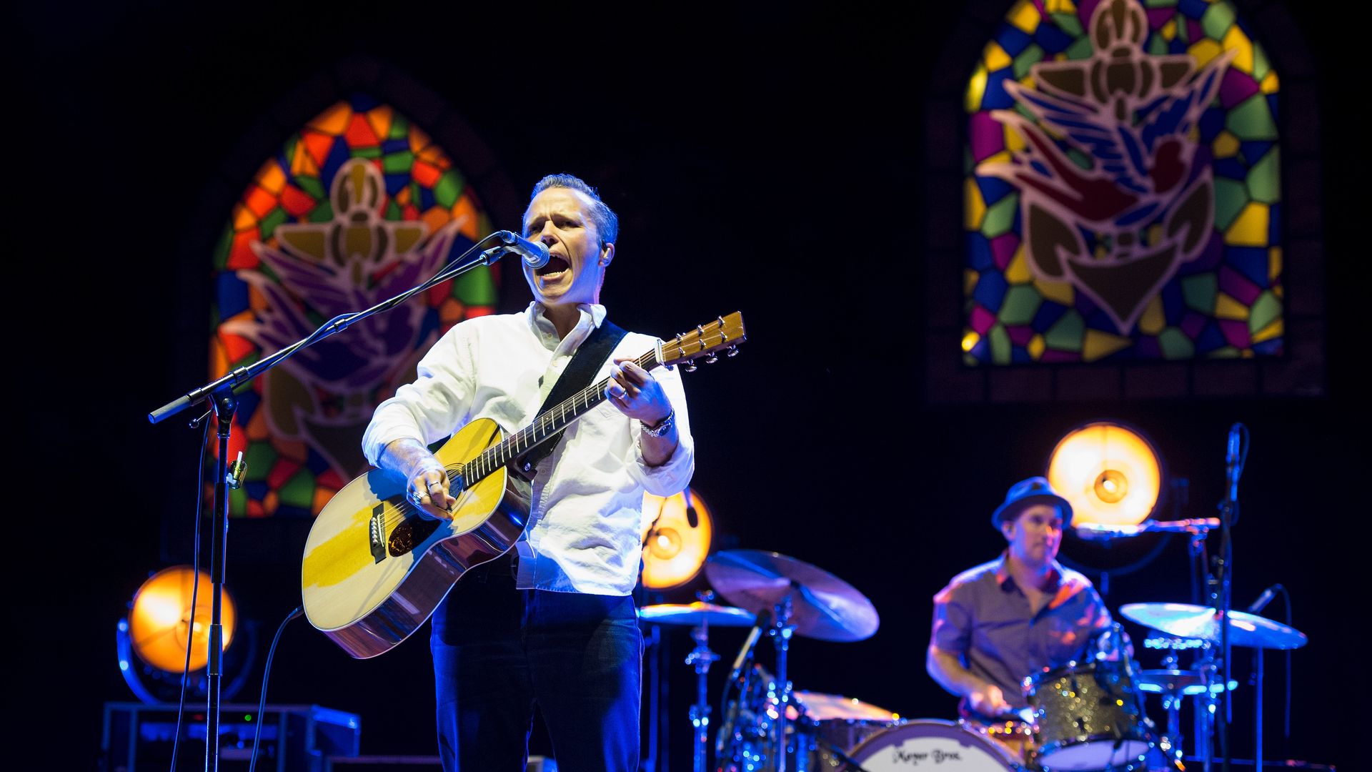 Male guitarist and vocalist in a white shirt sings into a microphone while playing an acoustic guitar on stage, with a drummer and colorful stained-glass windows in the background.