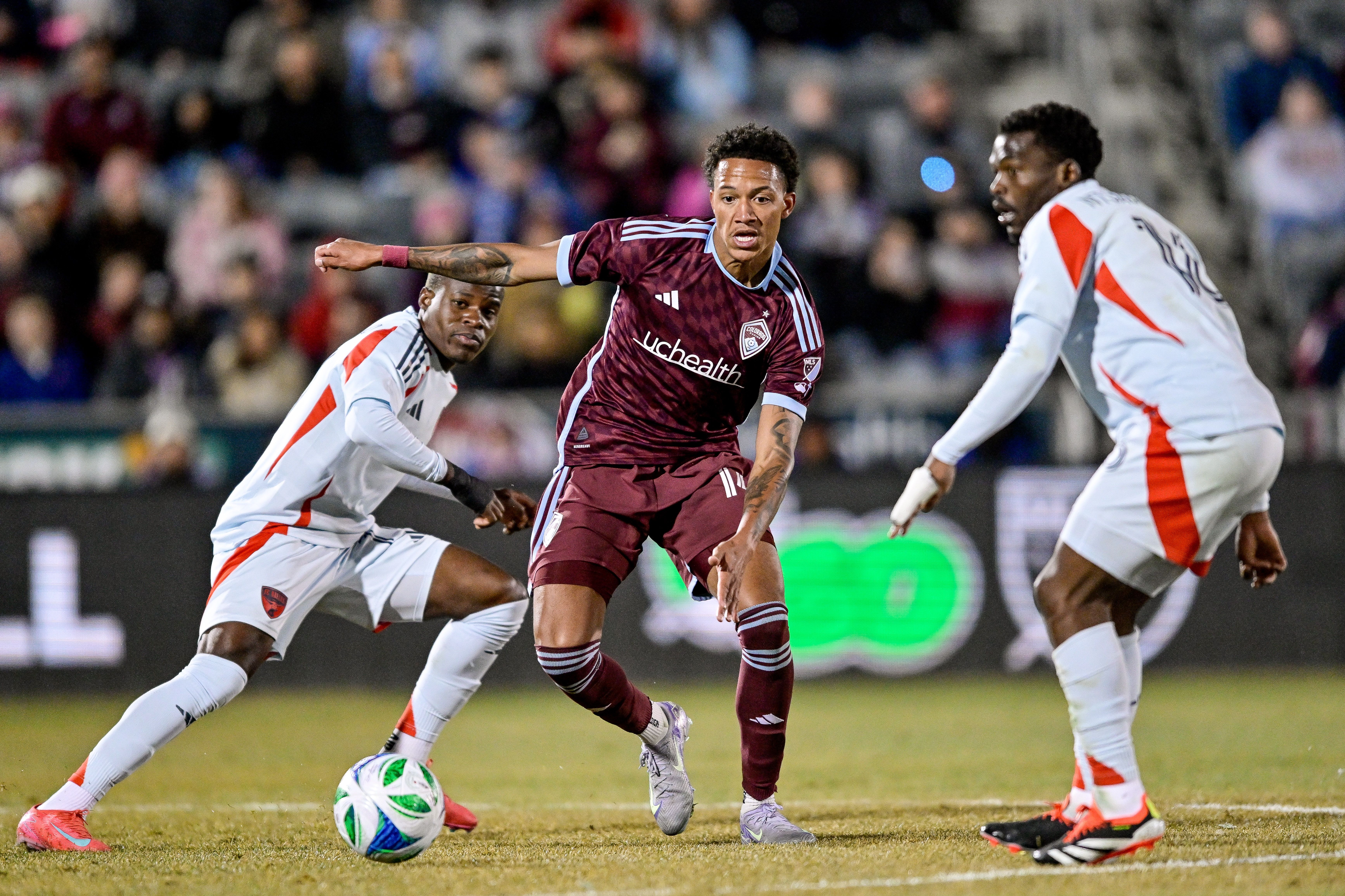 A soccer player in a burgundy jersey dribbles a ball between two players in white jerseys. 