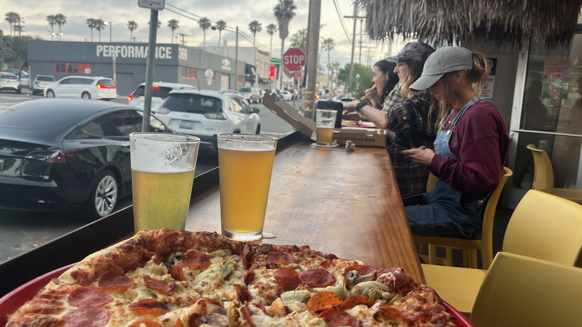 A pepperoni pizza and two beers sit on a ledge of a patio facing the street with palm trees in the background.