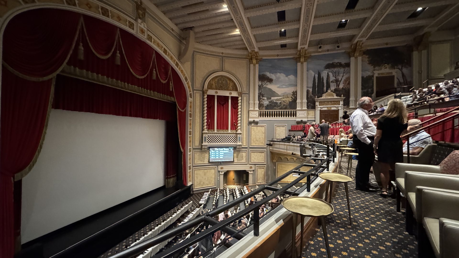 Interior view of a classic theater with red velvet curtains on stage, ornate walls with paintings, and audience seating including balcony tables with people mingling.
