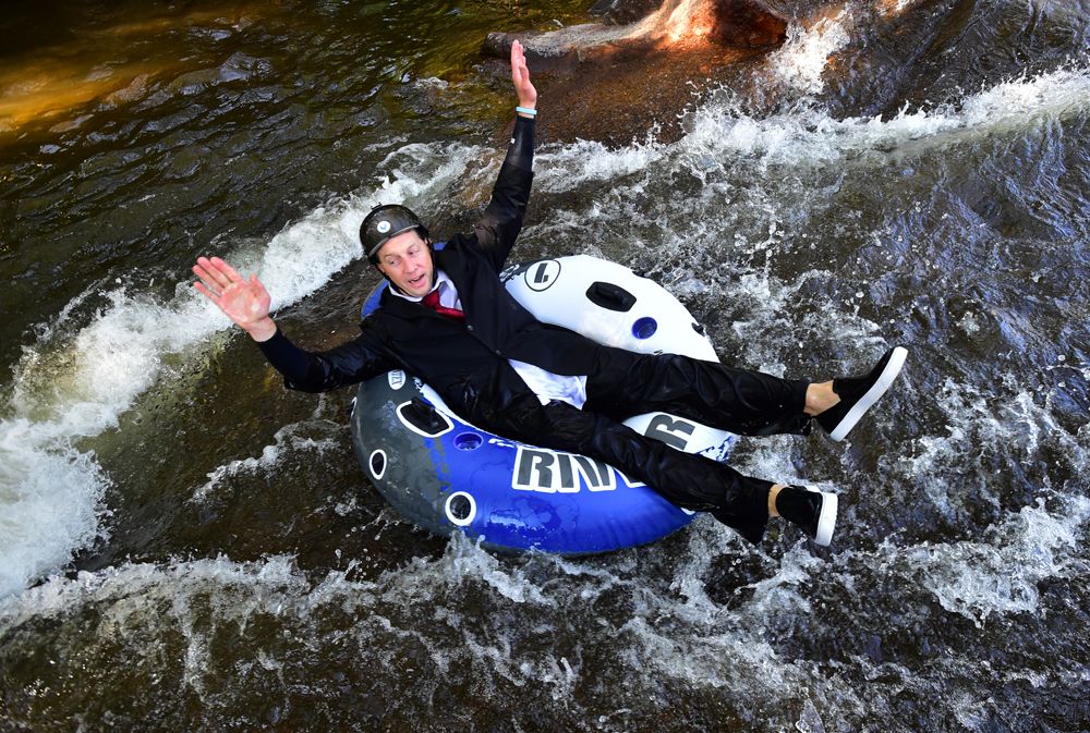 Man in a black suit, red tie, and helmet riding a blue and white inflatable tube down a fast-flowing river with white water rapids.
