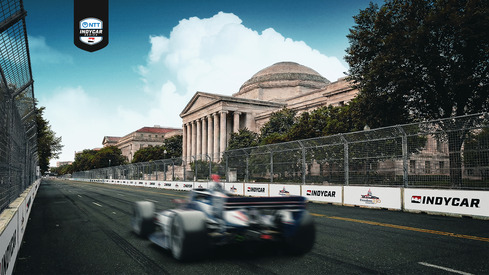 Blurred IndyCar racing past classical buildings with columns, surrounded by trees and fencing, under a blue sky with white clouds near NTT IndyCar Series signage.