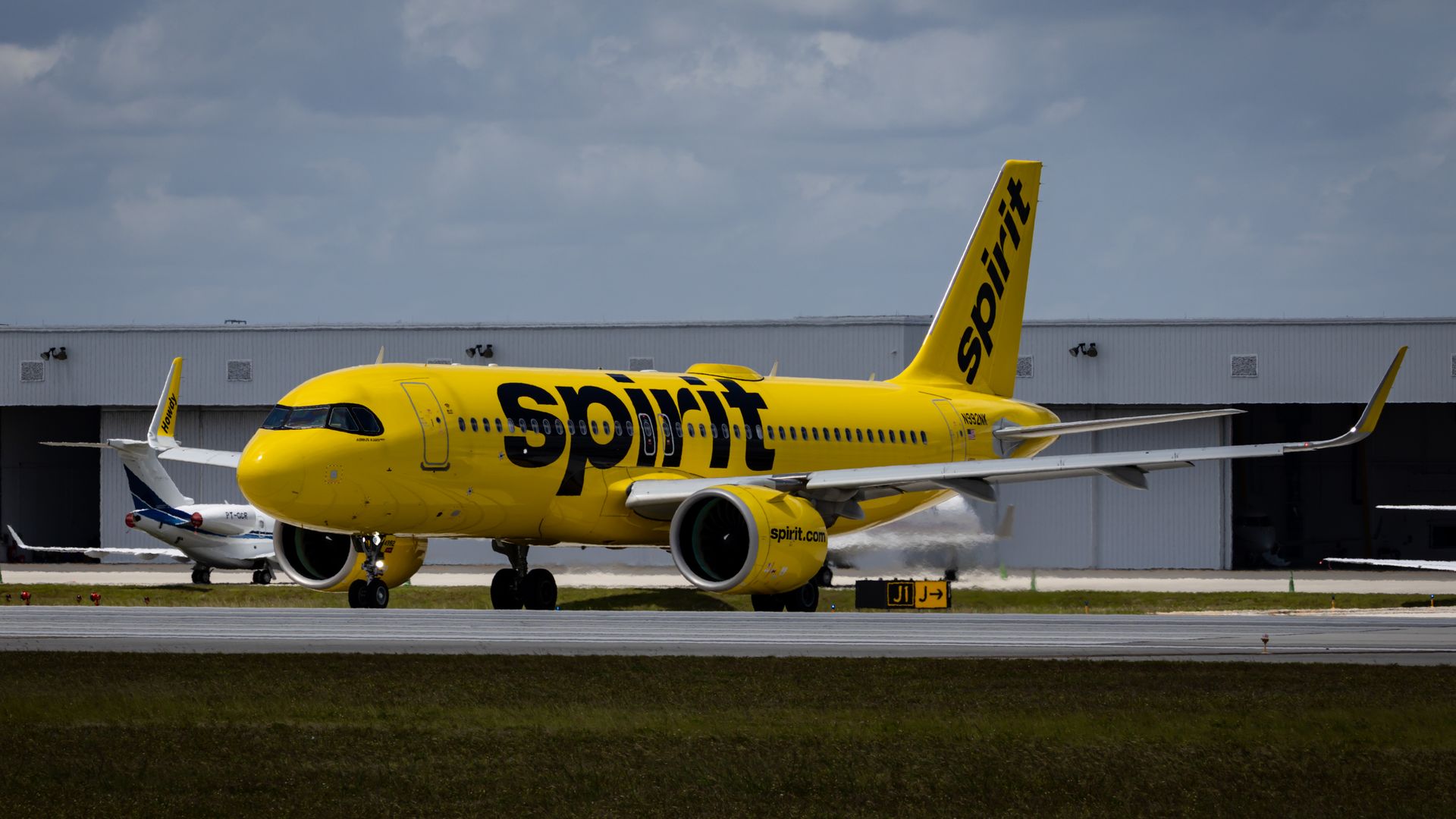 A Spirit Airlines plane, yellow with the word SPIRIT painted on the side, sits in front of an airport hangar. 