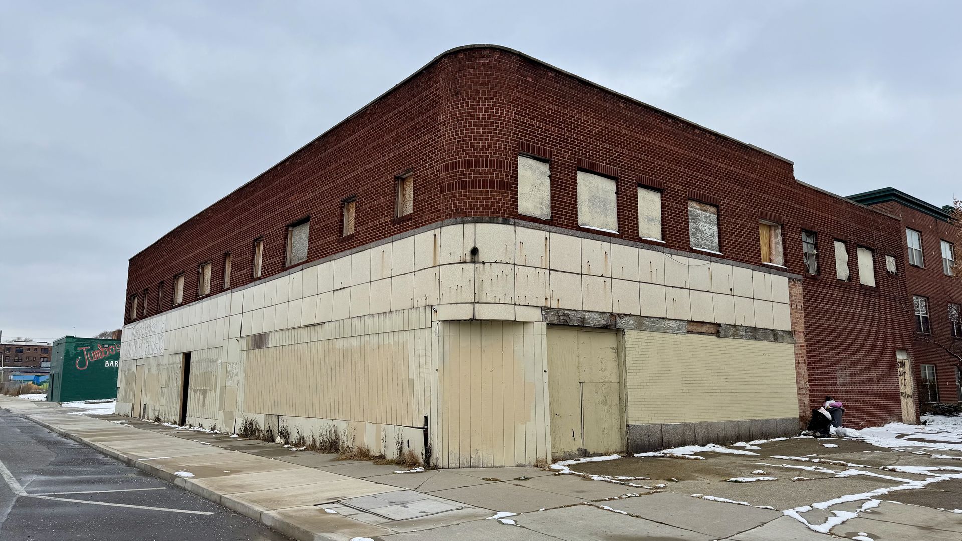 Corner view of a two-story, boarded-up brick building with faded beige panels and snow patches on the sidewalk and parking lot under a cloudy gray sky.
