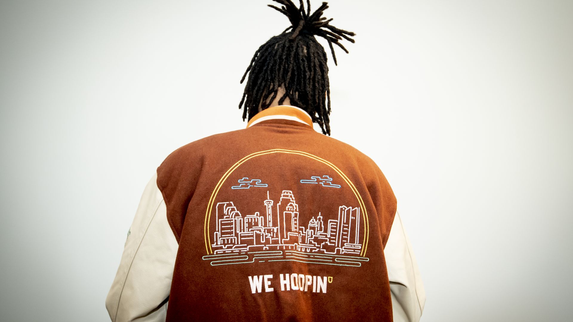 Devin Vassell facing away from the camera, showing the back of his letterman-style jacket, which features the San Antonio skyline with the words "we hoopin'." 