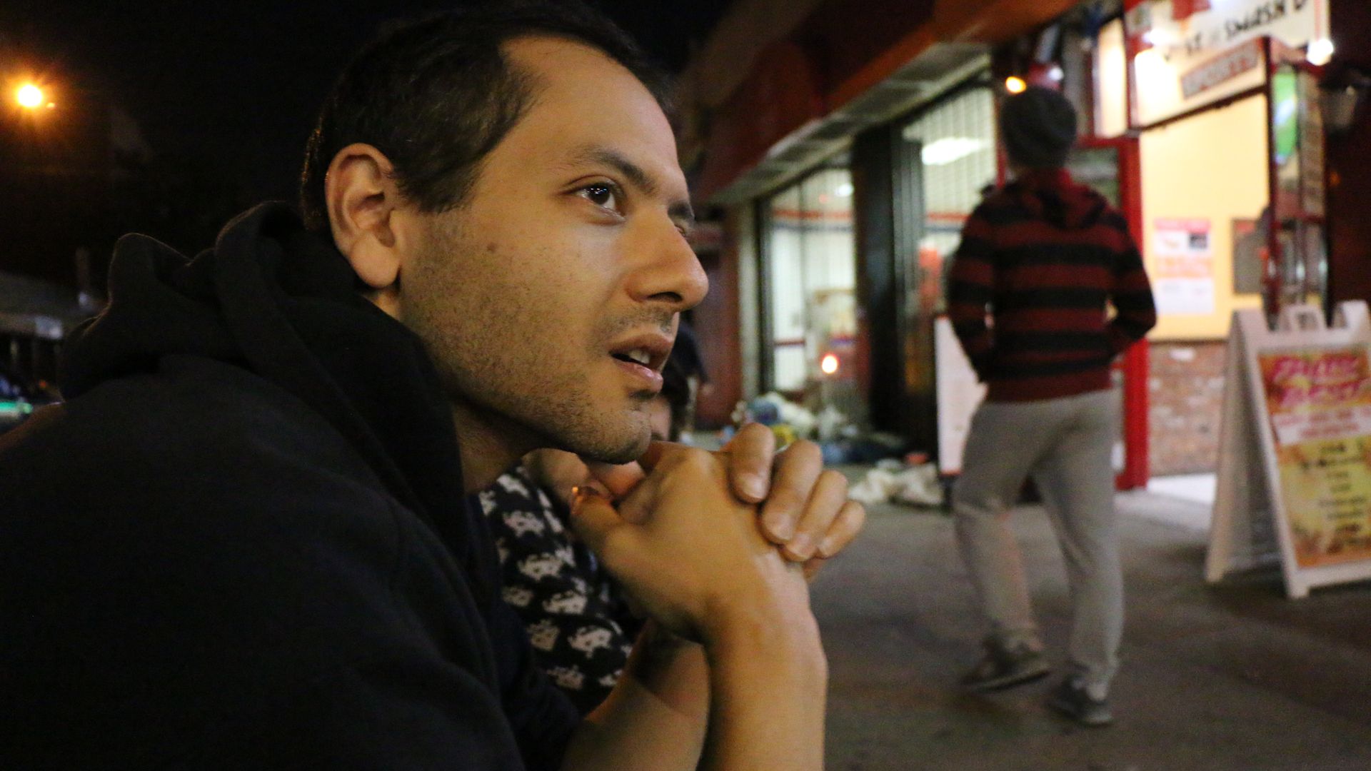 A man in profile, sitting with his chin on his hands, looking to one side,  in the evening by a few lit shops.